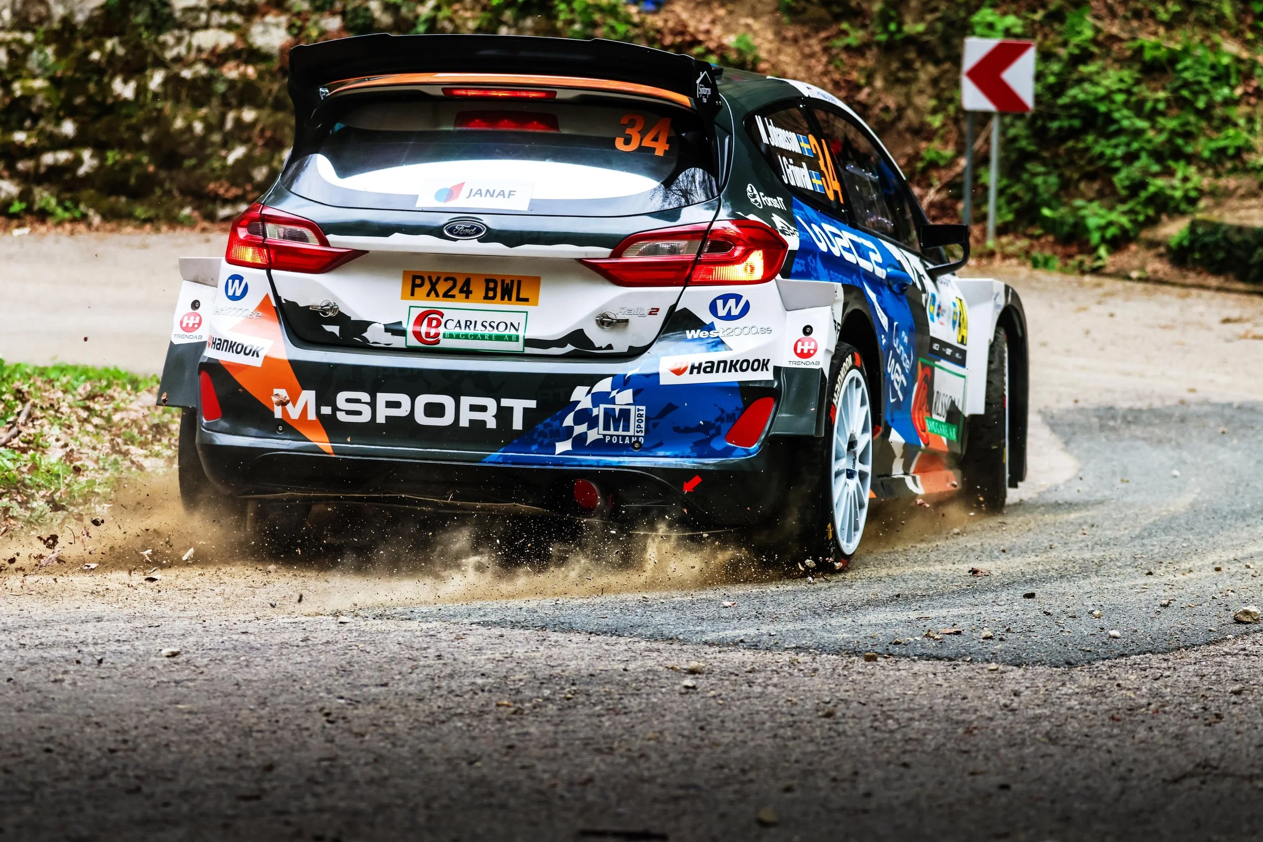 A rally car racing on an unpaved road, kicking up dust and debris, surrounded by trees and a wooden sign with a red arrow.