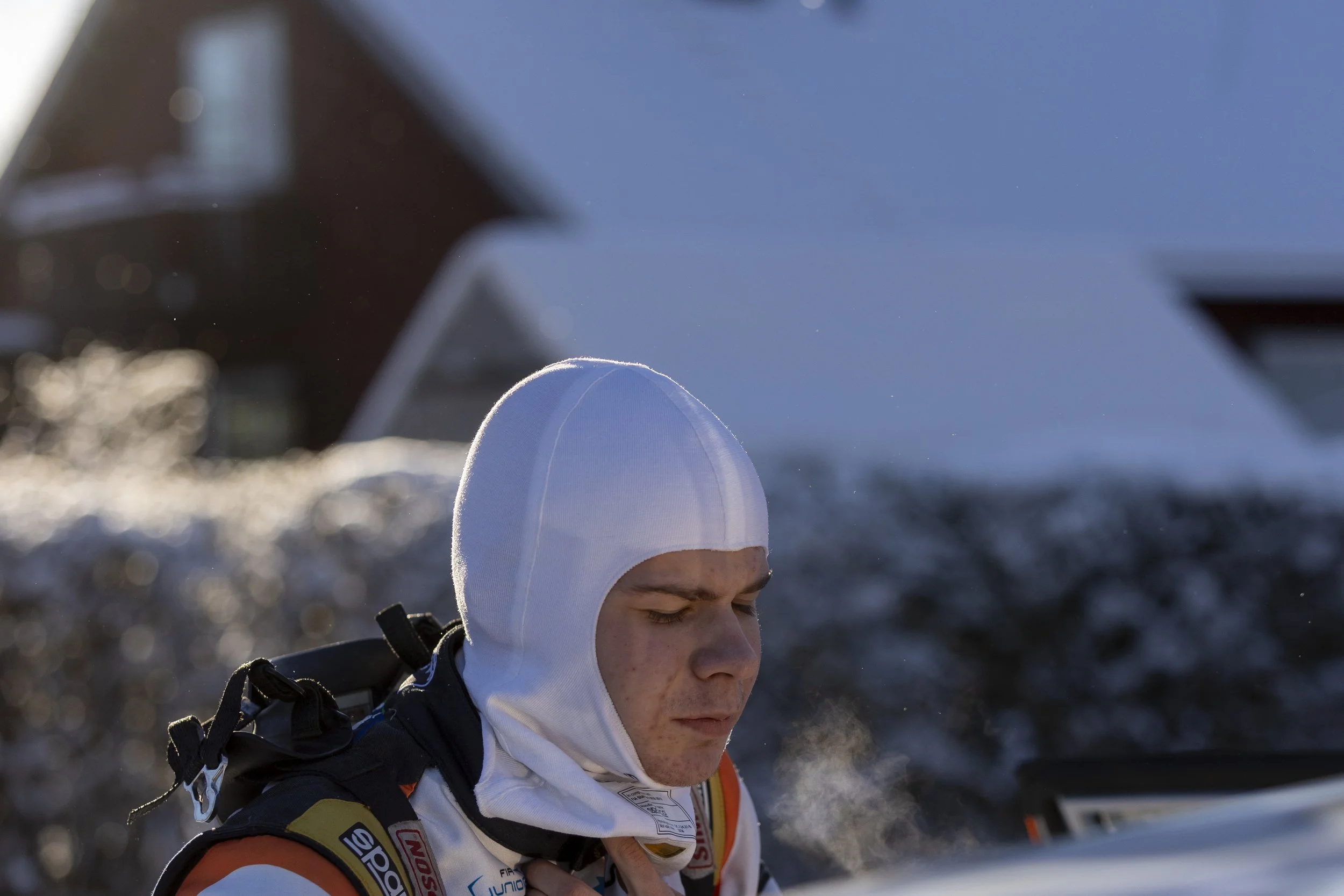 A person with light skin wearing a white hood and black racing gear, standing outdoors with snow, with mountains and blue sky in the background.