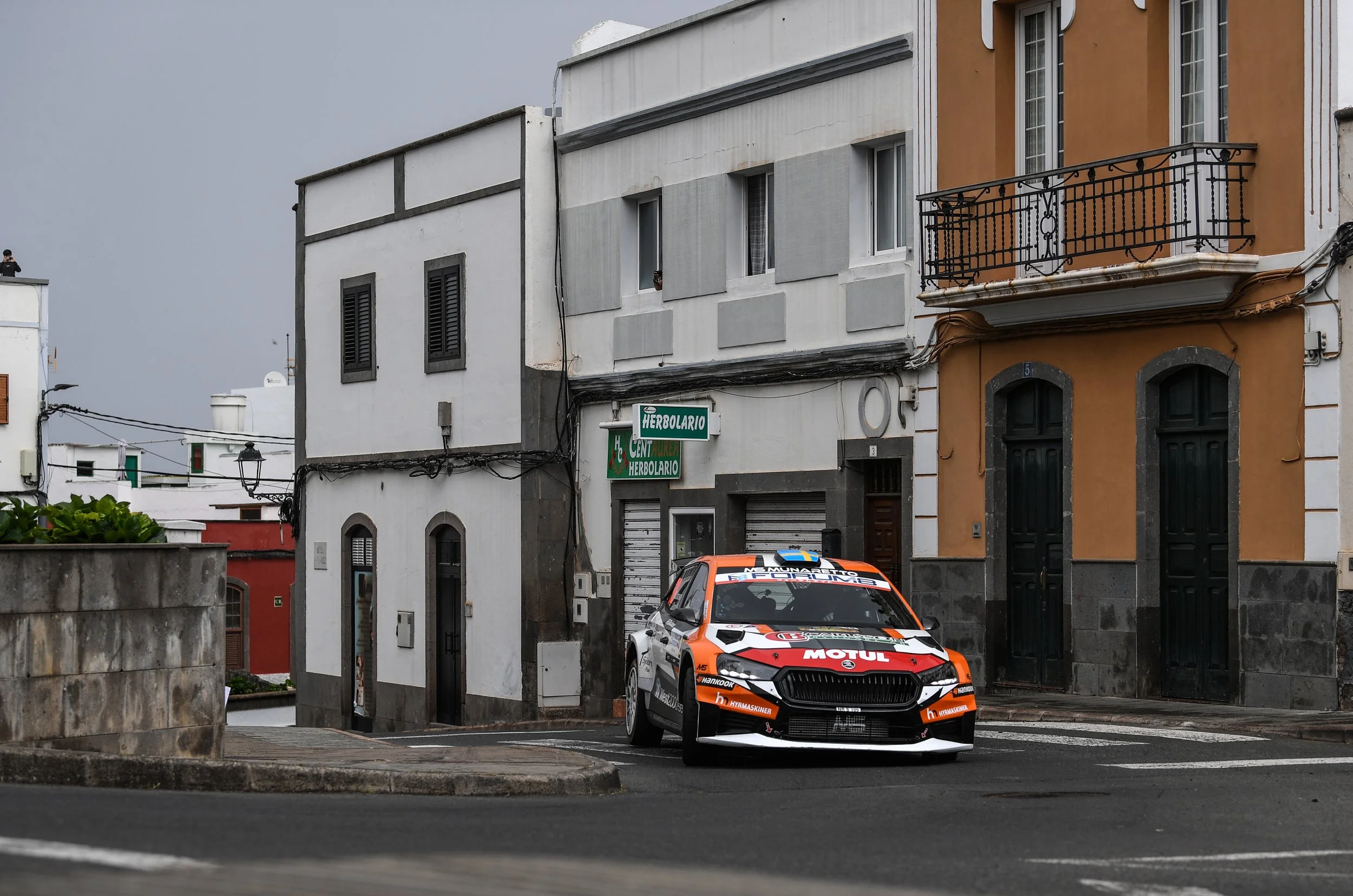 A rally car with sponsors' logos parked on a street in front of European style buildings, with one building painted orange and another painted white with gray accents.