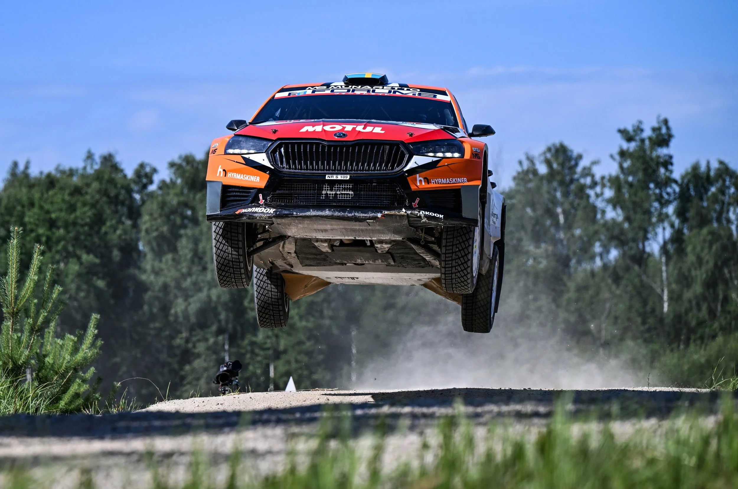 A race car in mid-air during a rally race, with trees in the background and the car flying above the dirt road.