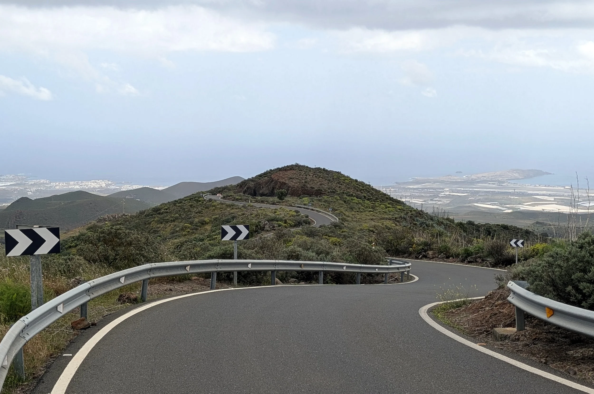 A winding mountain road with guardrails and black-and-white chevron road signs, overlooking hills, water, and buildings in the distance under a cloudy sky.