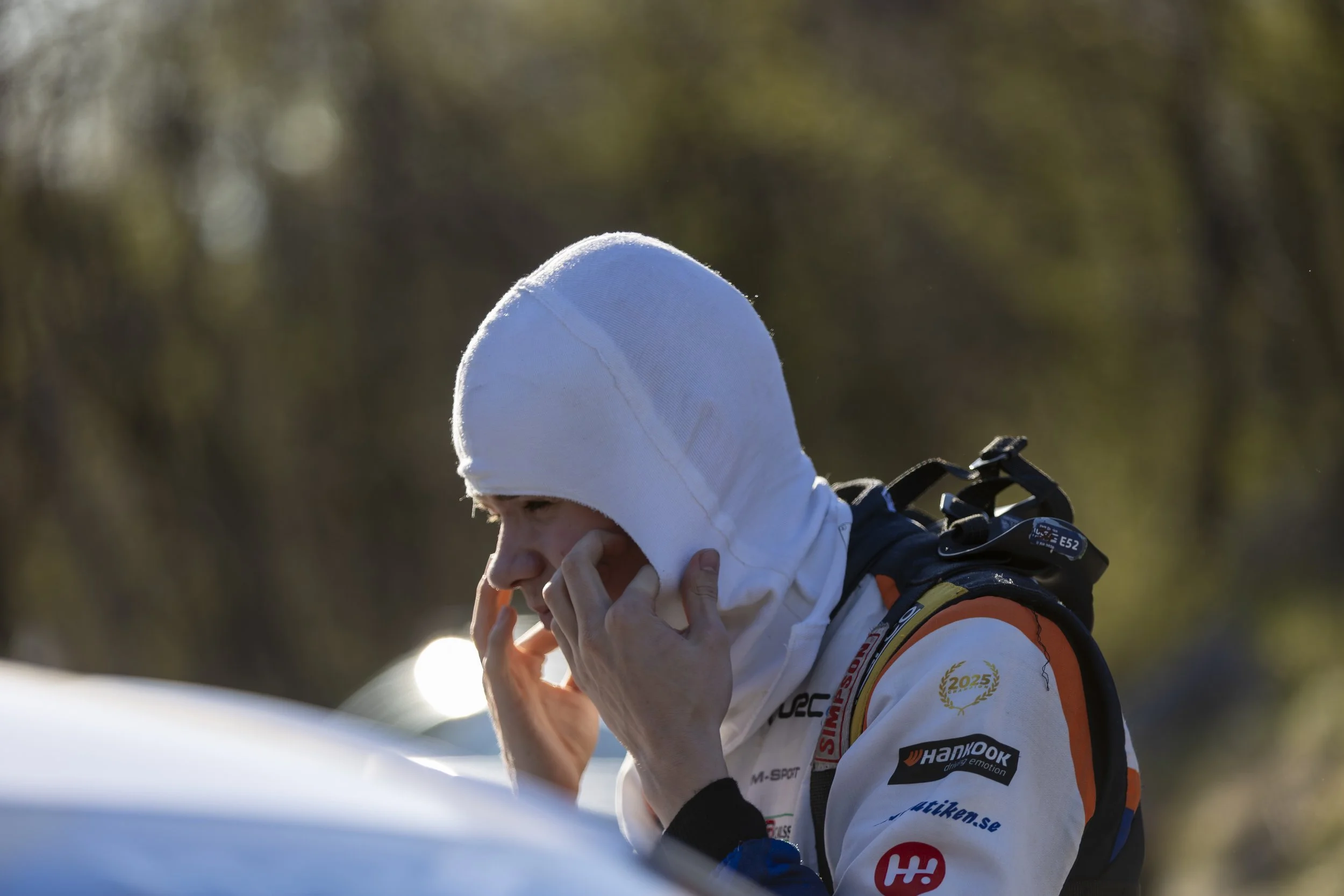 Person wearing a white hood and white racing suit with sponsor logos, holding their face with one hand, in an outdoor setting.