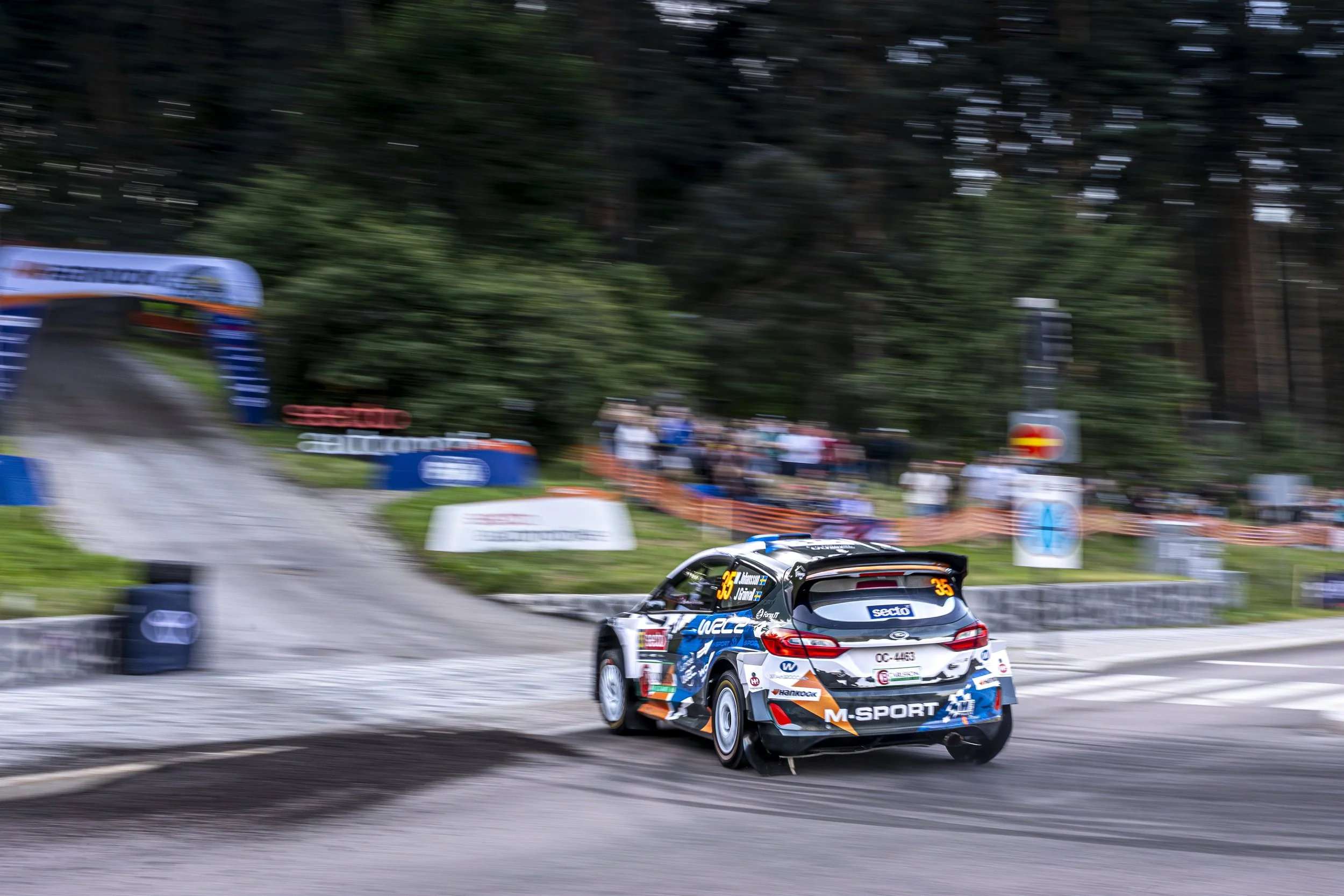 A rally car racing on a dirt track with trees in the background, showing motion blur and speed.