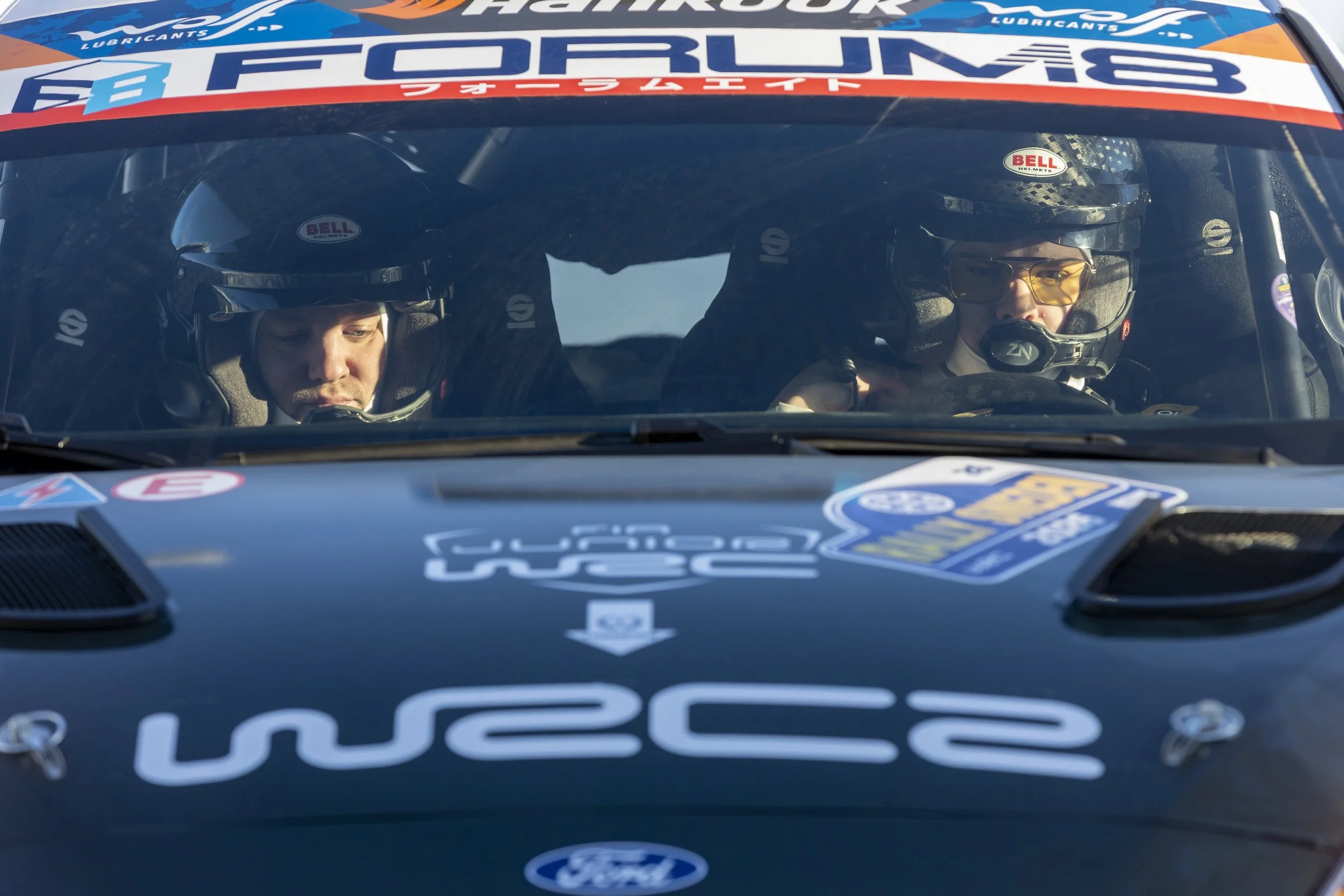 Close-up of two race car drivers inside a race car, both wearing black helmets with Bell logos, and racing suits. The dashboard and interior of the car are visible, with sponsorship stickers and decals on the car's windshield and hood.
