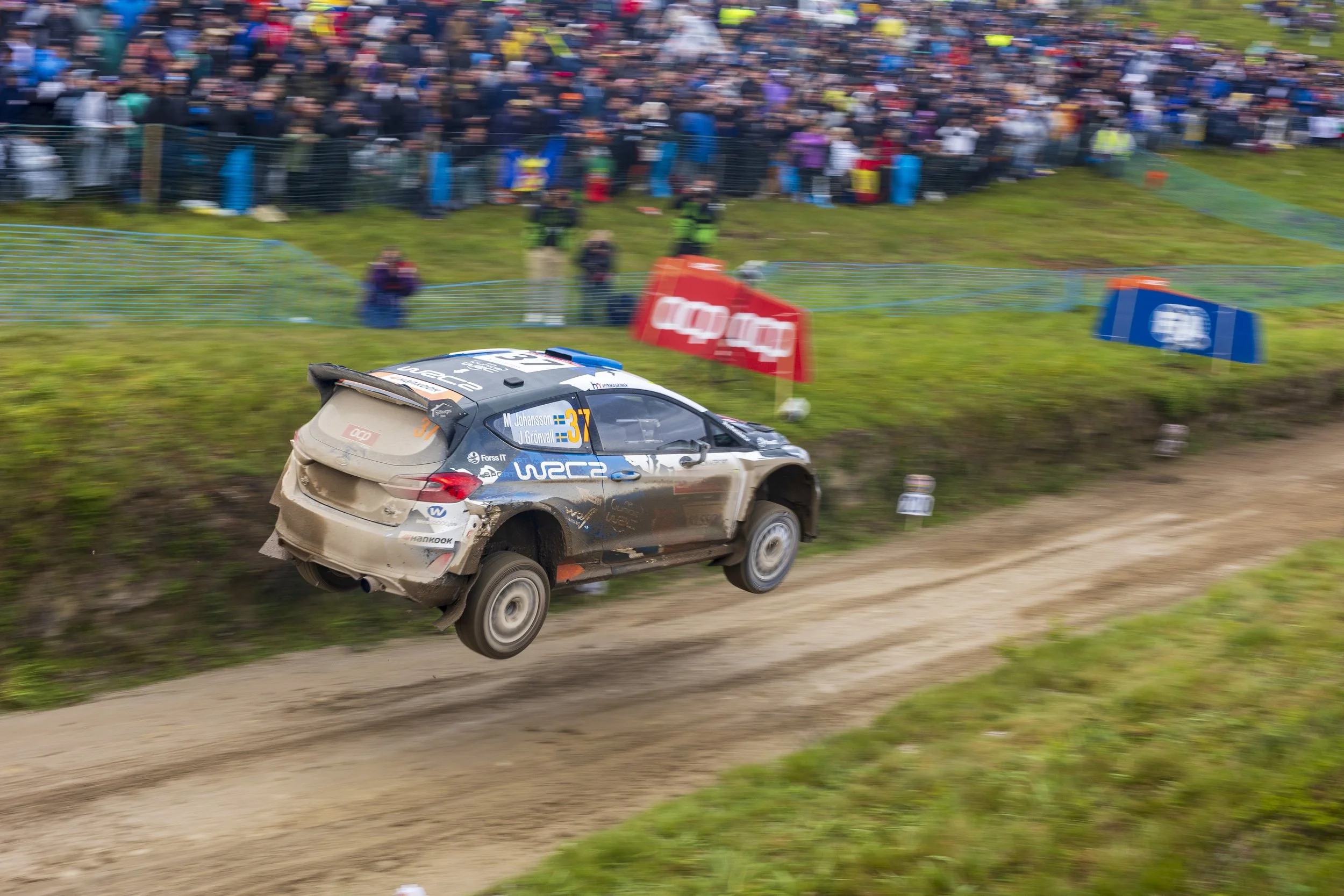 A rally car with the number 37 speeding on a dirt track with a large crowd of spectators watching from behind a fence.