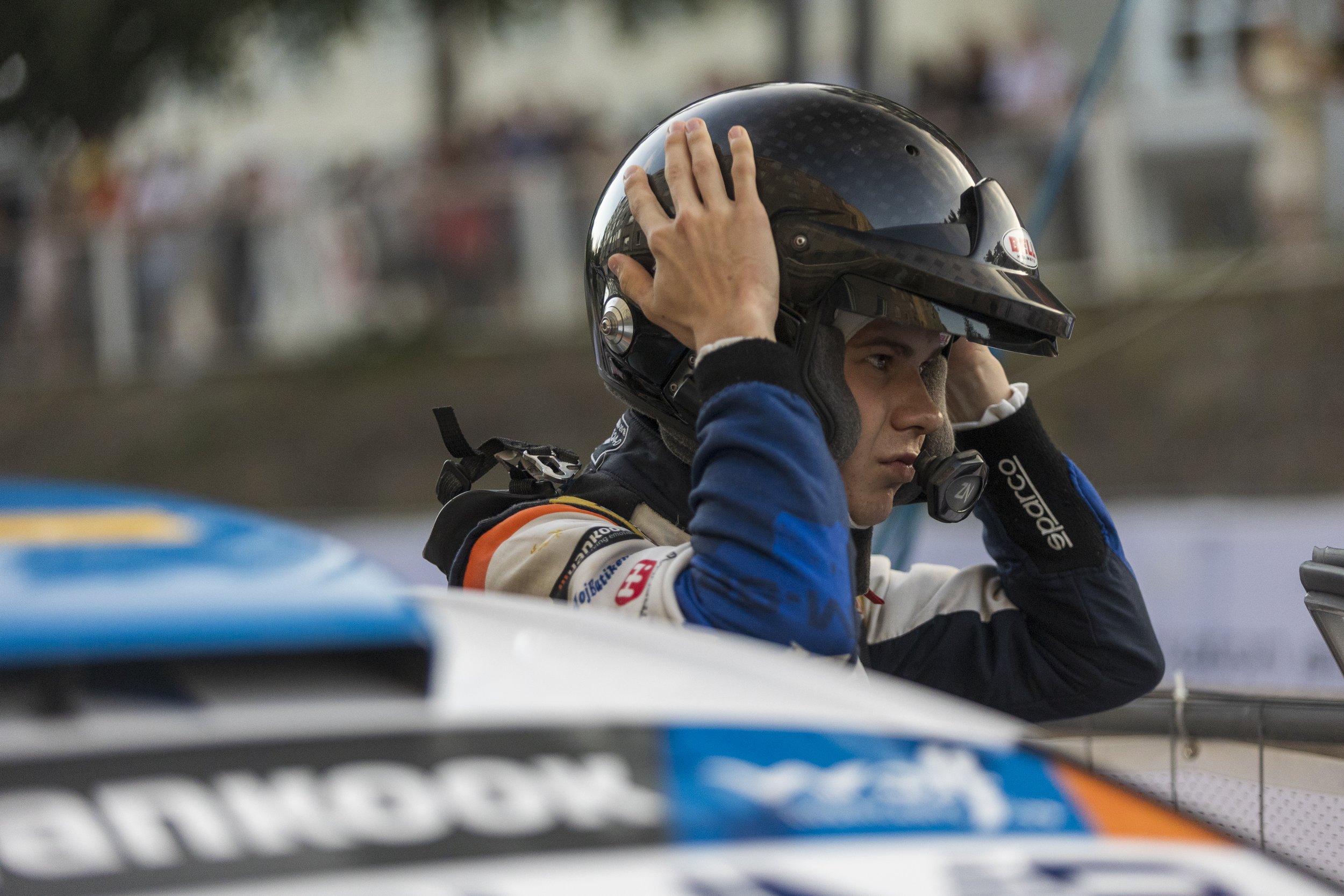 A race car driver in a racing suit adjusts a black motorcycle helmet with a reflective visor.