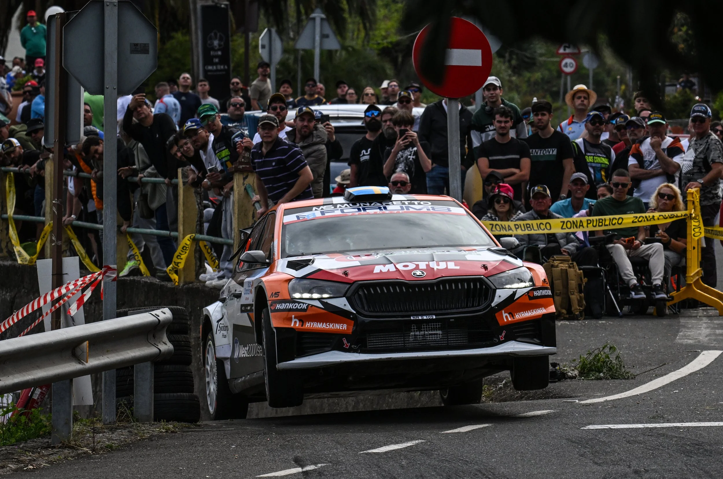 A rally car driving on a road with a crowd of spectators behind a barrier watching the race. The crowd is standing behind yellow caution tape marked 'Zona Publico'. Vehicle branding includes 'MS Munarieto' and logos of sponsors like Hankook and Hyund