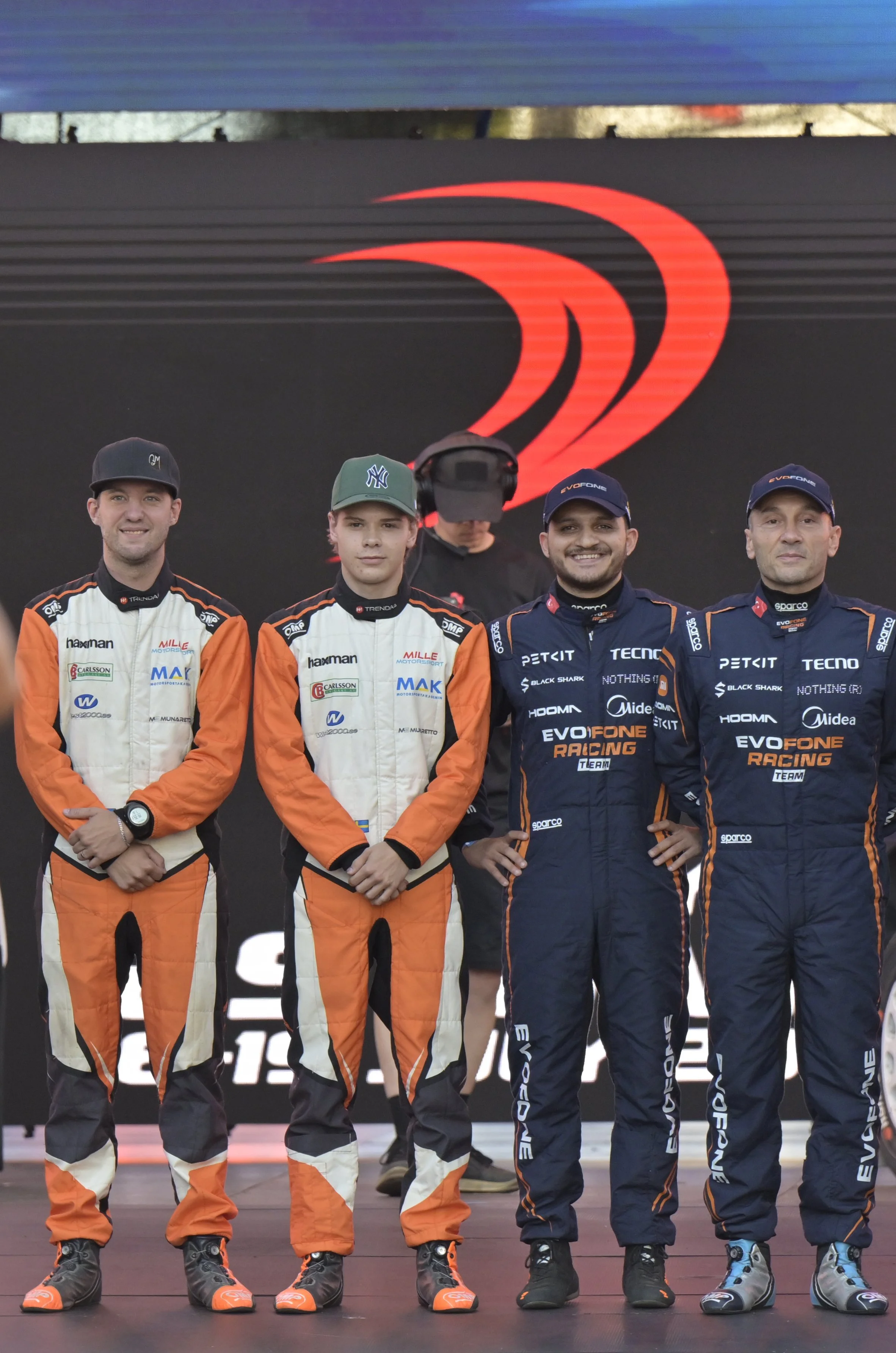 Five race car drivers standing in a line, wearing racing suits and caps, in front of a backdrop with a stylized red logo.
