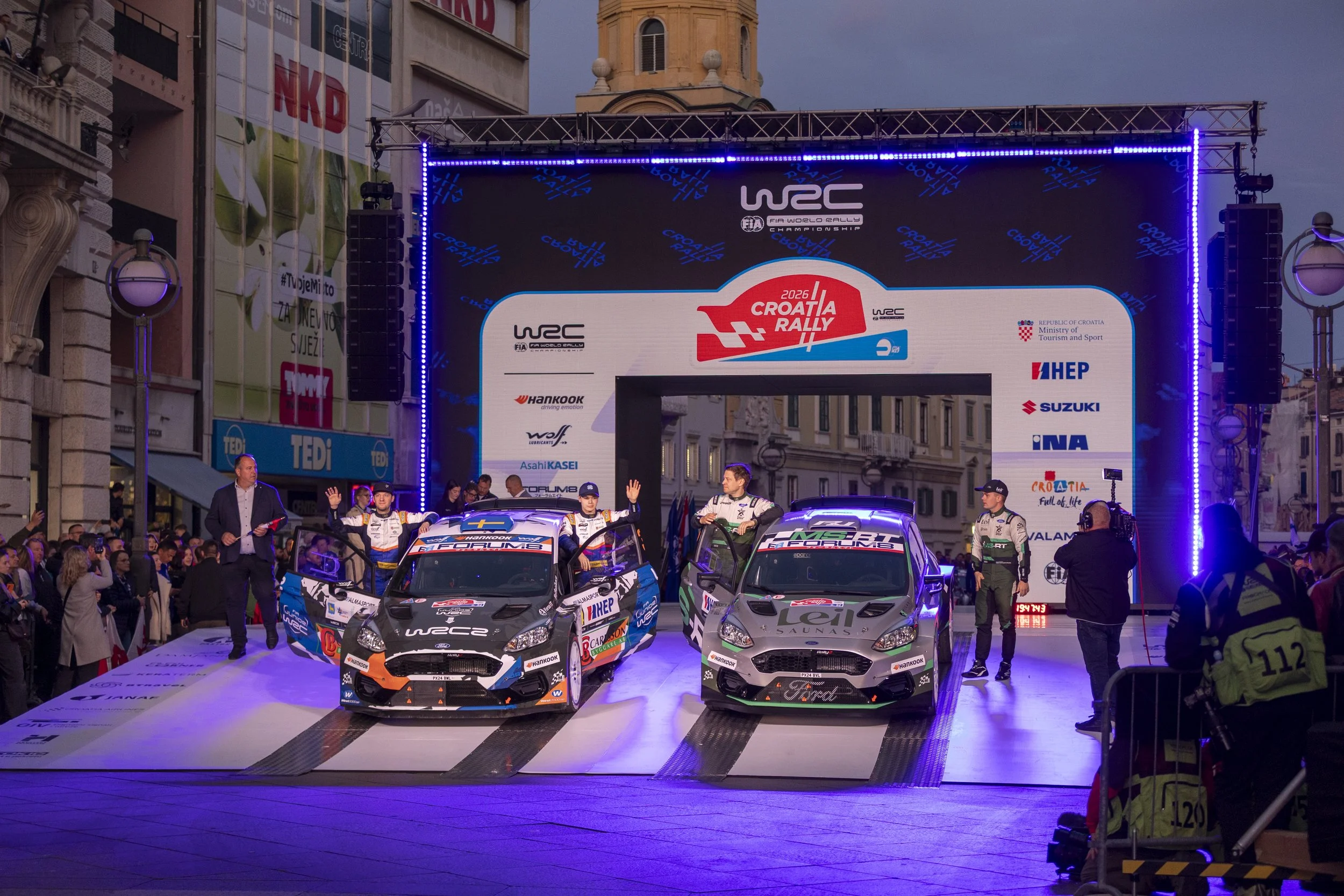Race cars and drivers on a podium during a rally event, with a large screen displaying event logos and sponsors in the background, and people surrounding the cars.