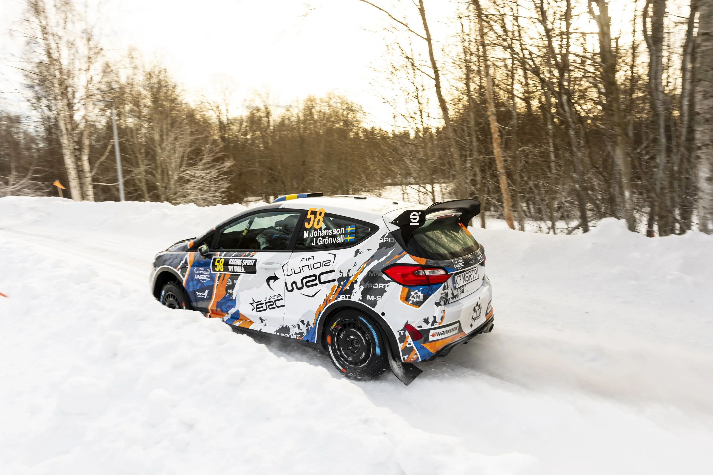 A rally car racing on a snow-covered road surrounded by leafless trees during winter, with the car displaying the number 58 and driver names M Johansson and J Grønval.