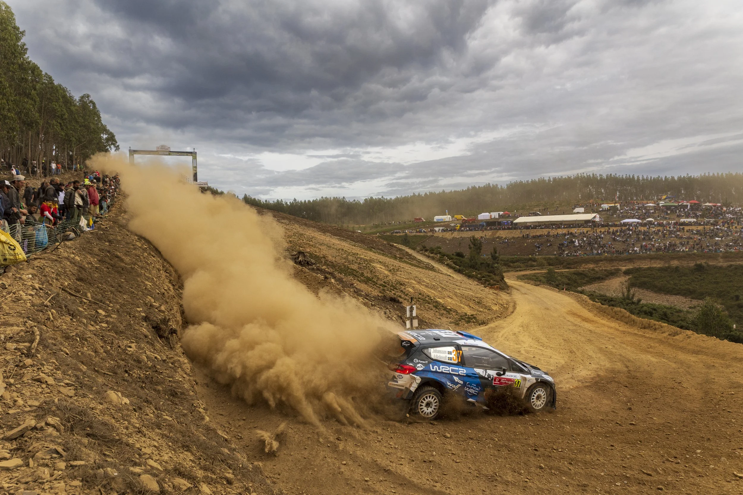 A rally car racing on a dirt track, kicking up a cloud of dust, with a large crowd of spectators on the hillside and in tents watching under a cloudy sky.