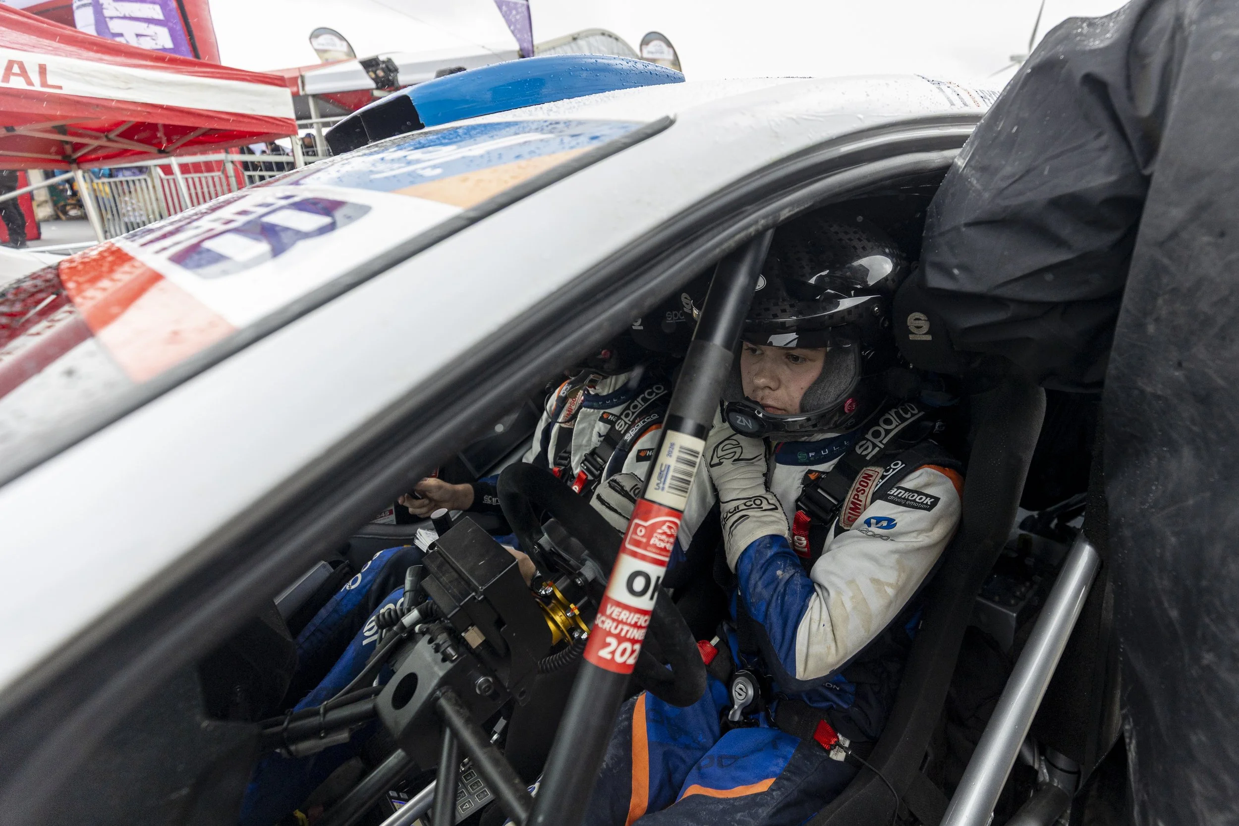 Race car driver wearing a helmet and racing suit inside a race car, preparing for a race, with pit crew working outside the car.