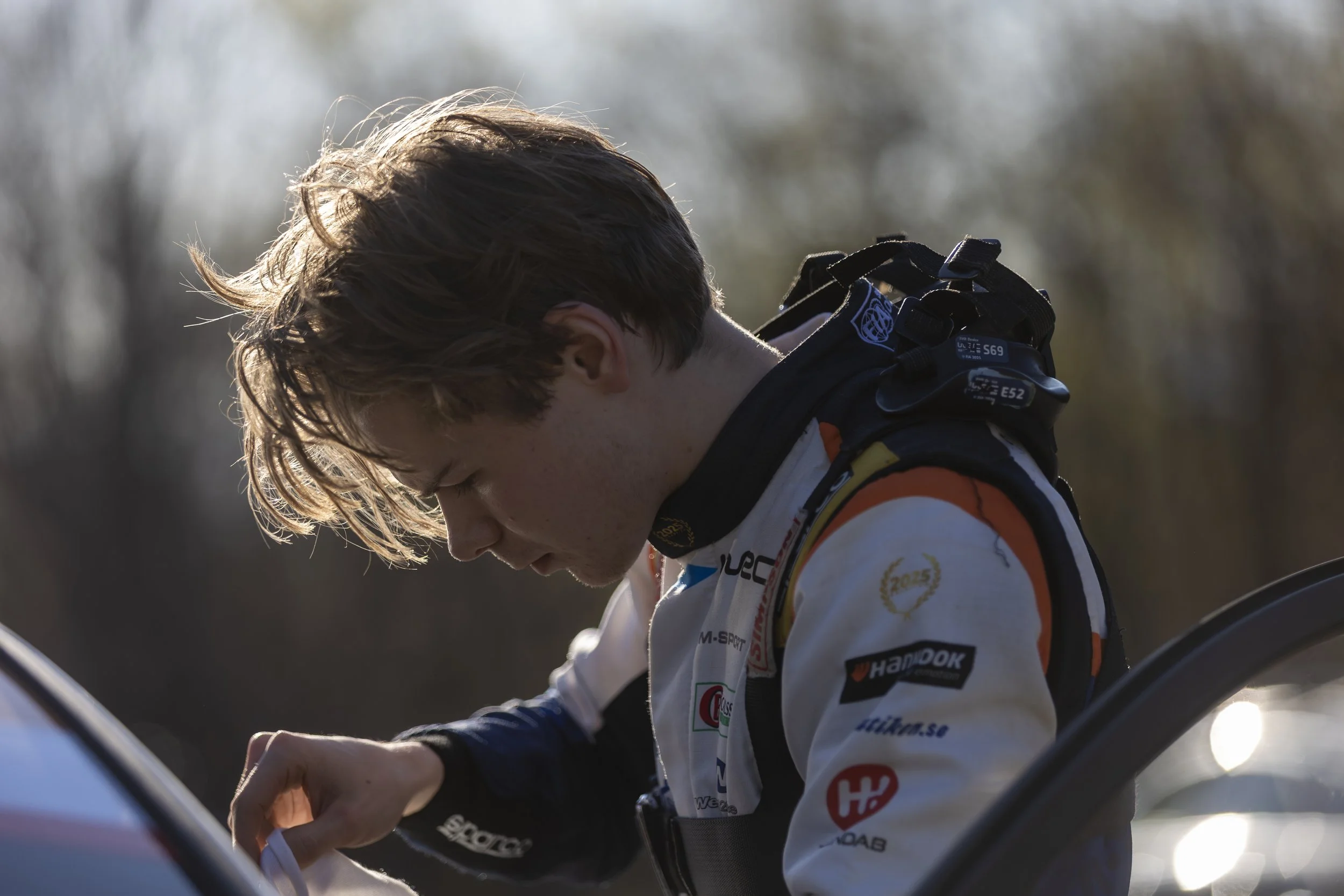 Close-up of a young male race car driver with brown hair, wearing a racing suit with logos, adjusting his helmet strap outdoors during daytime.