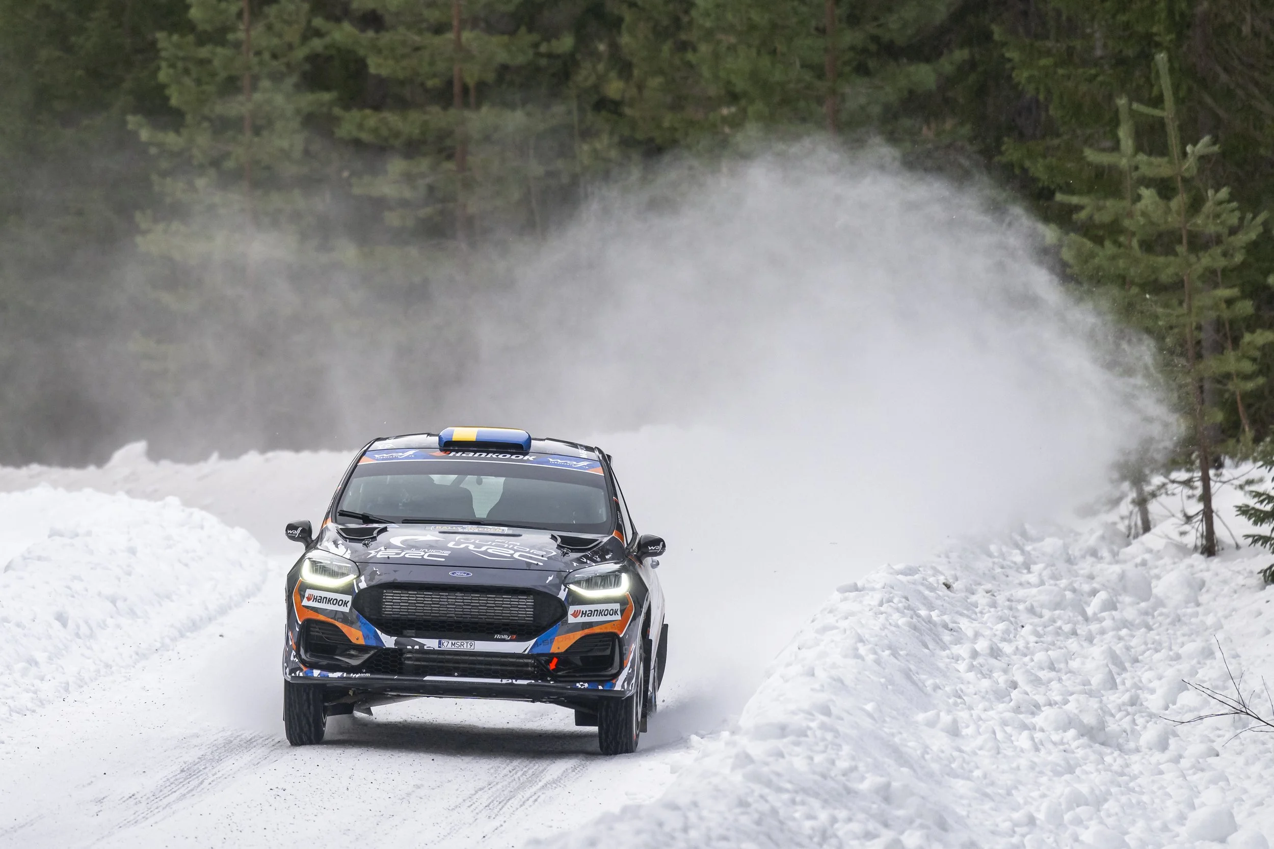 A rally car driving on a snowy road in a forest, with steam or smoke rising behind it.