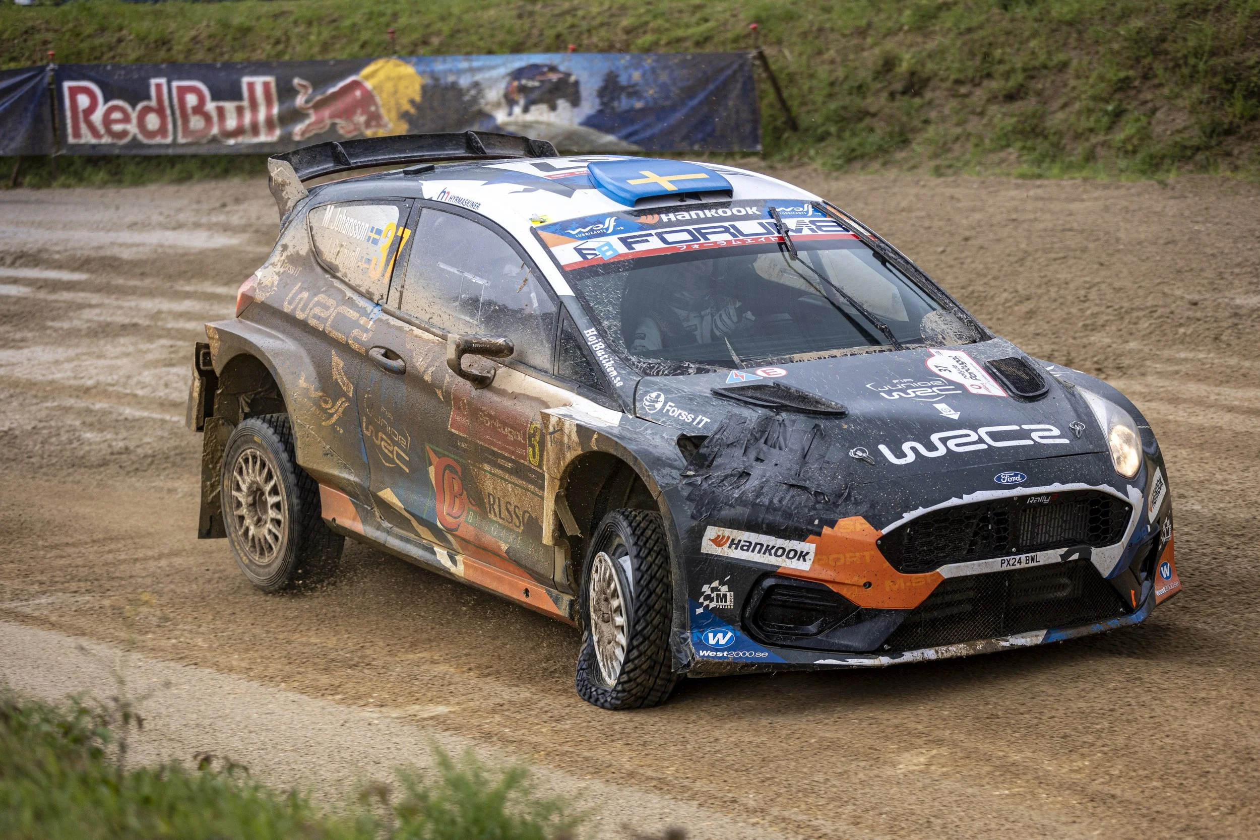 A rally car racing on a dirt track, kicking up mud and dirt, with sponsor logos and Swedish flags on the vehicle. A Red Bull banner is displayed in the background.