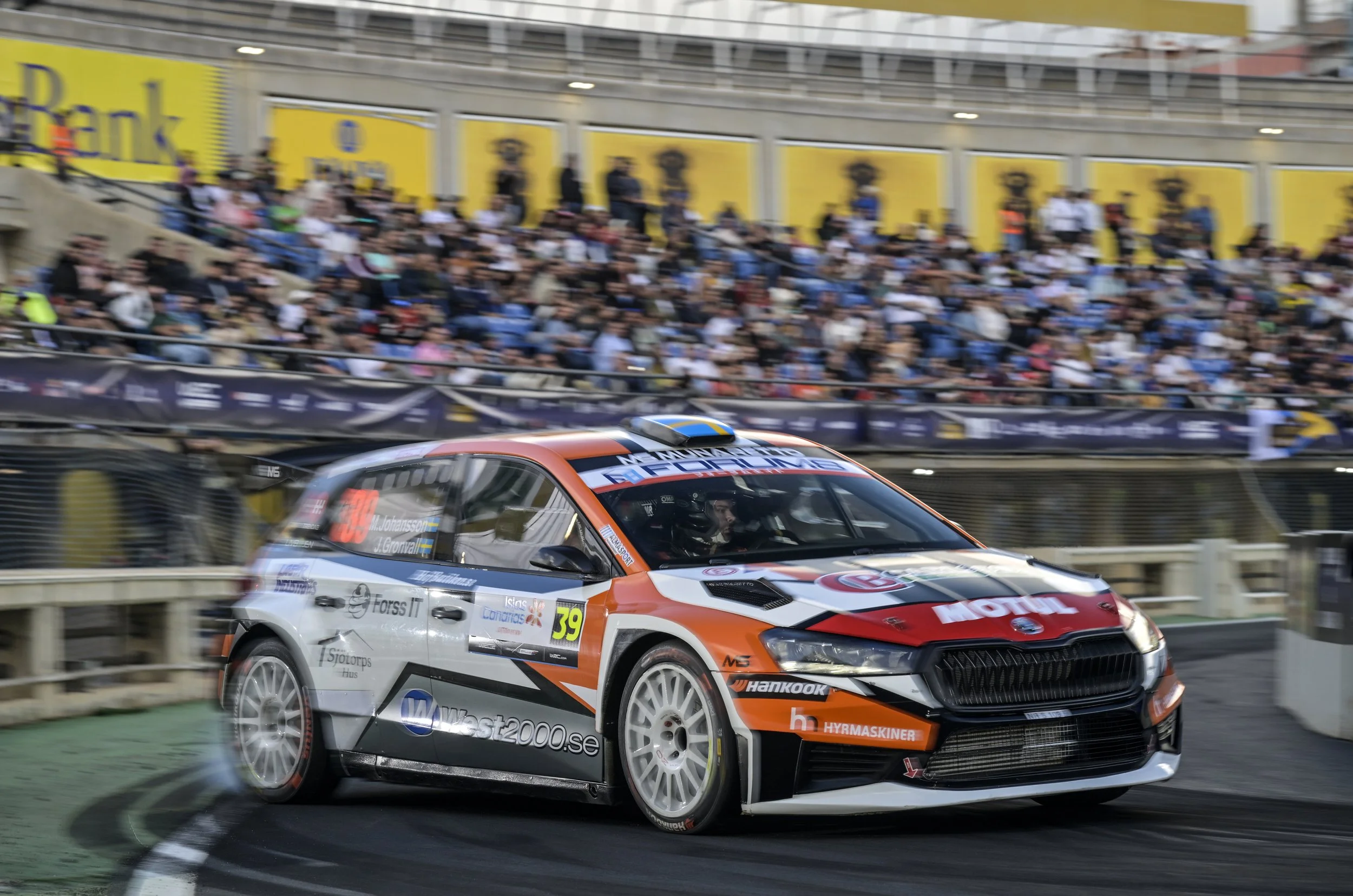 A race car with number 39 and sponsorship logos speeding on a race track, with a blurred crowd in the grandstands and advertising banners in the background.