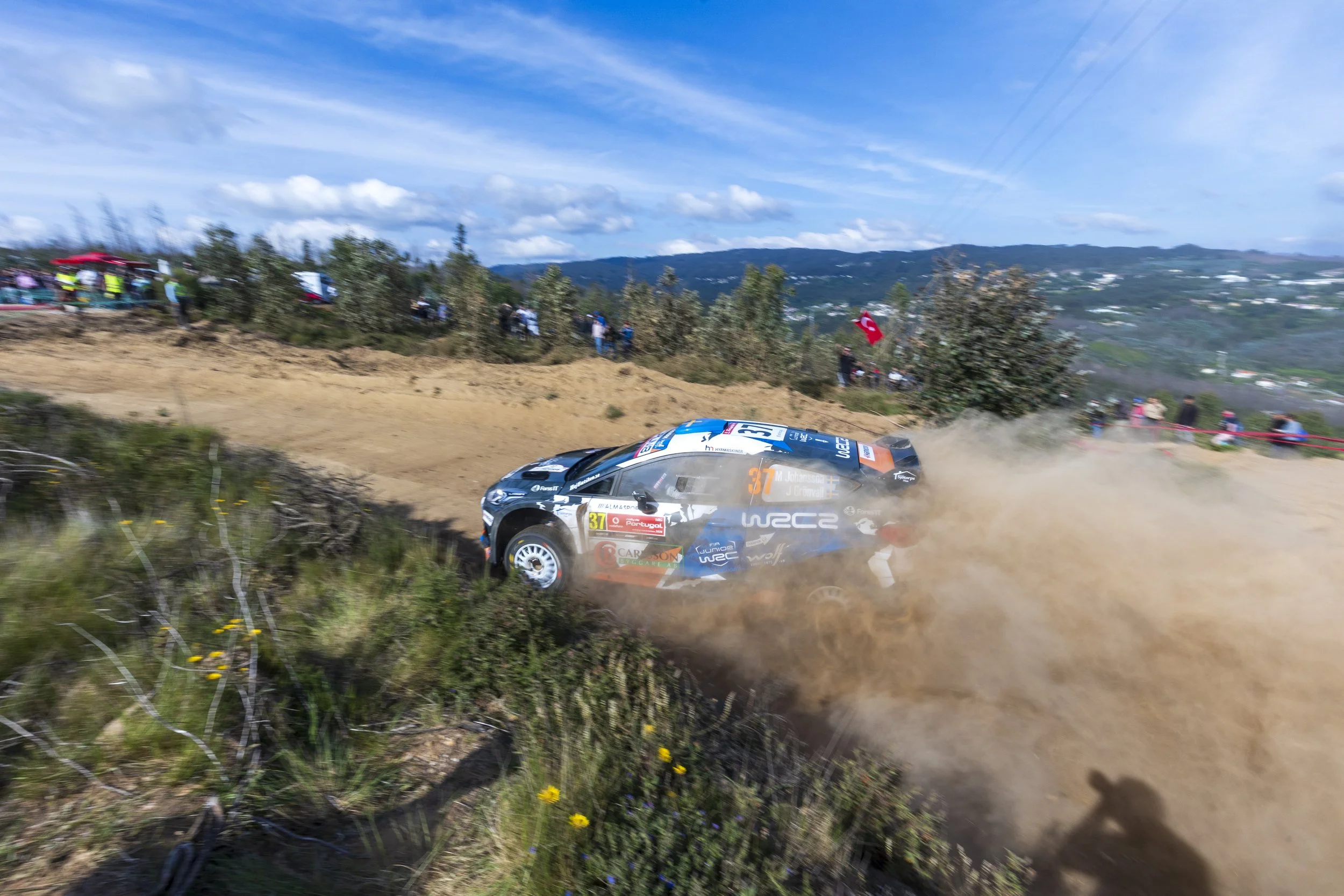 A rally car speeding along a dirt road, kicking up dust, with spectators watching from the hillside. The landscape features distant hills and a partly cloudy sky, with a Turkish flag visible among the spectators.