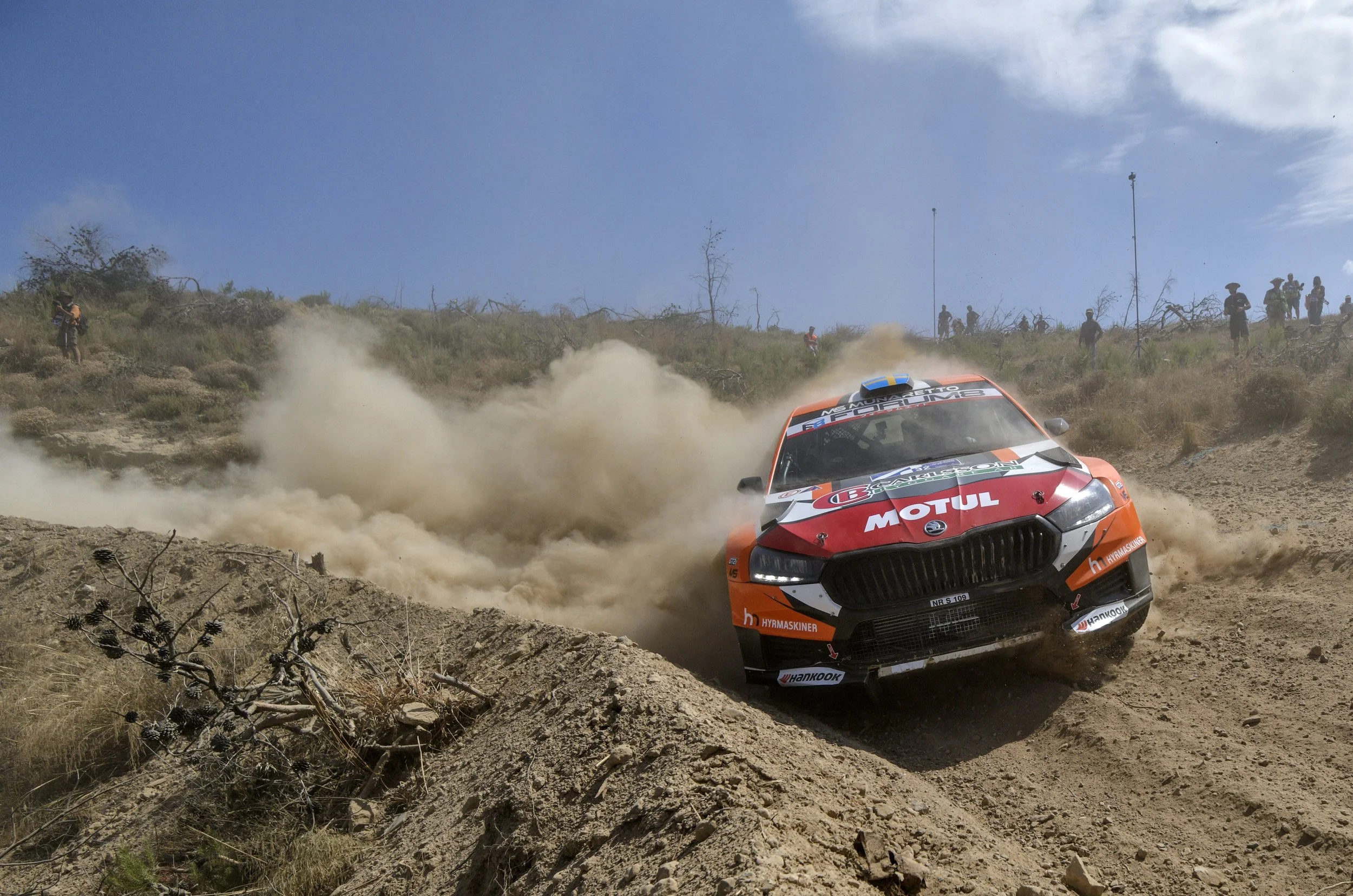 An orange rally car kicking up dust as it races on a rugged dirt trail, with spectators and team members watching from a hillside in the background.