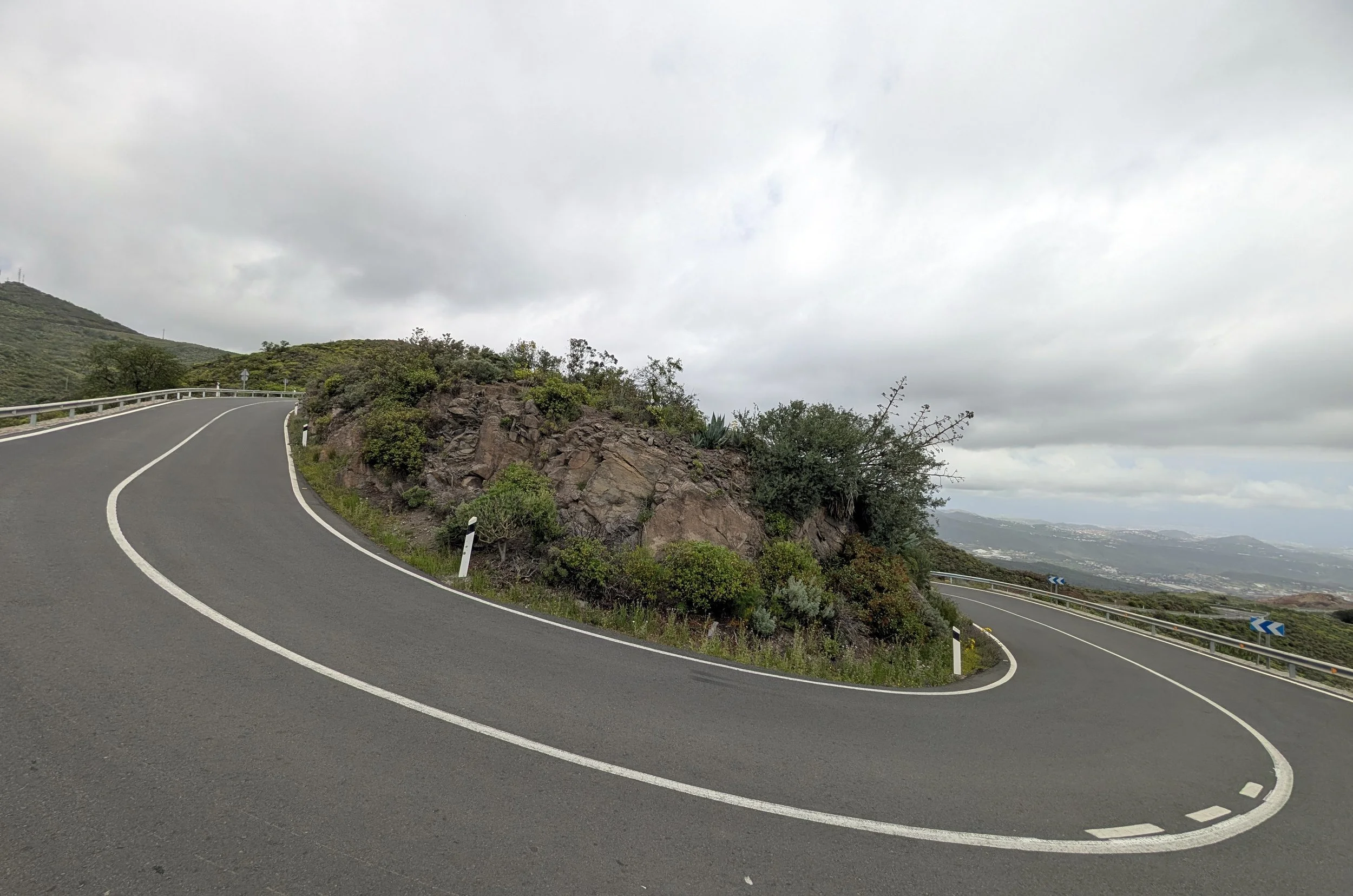 A winding mountain road with guardrails, rock formations, and green shrubbery, under a cloudy sky in a hilly landscape.