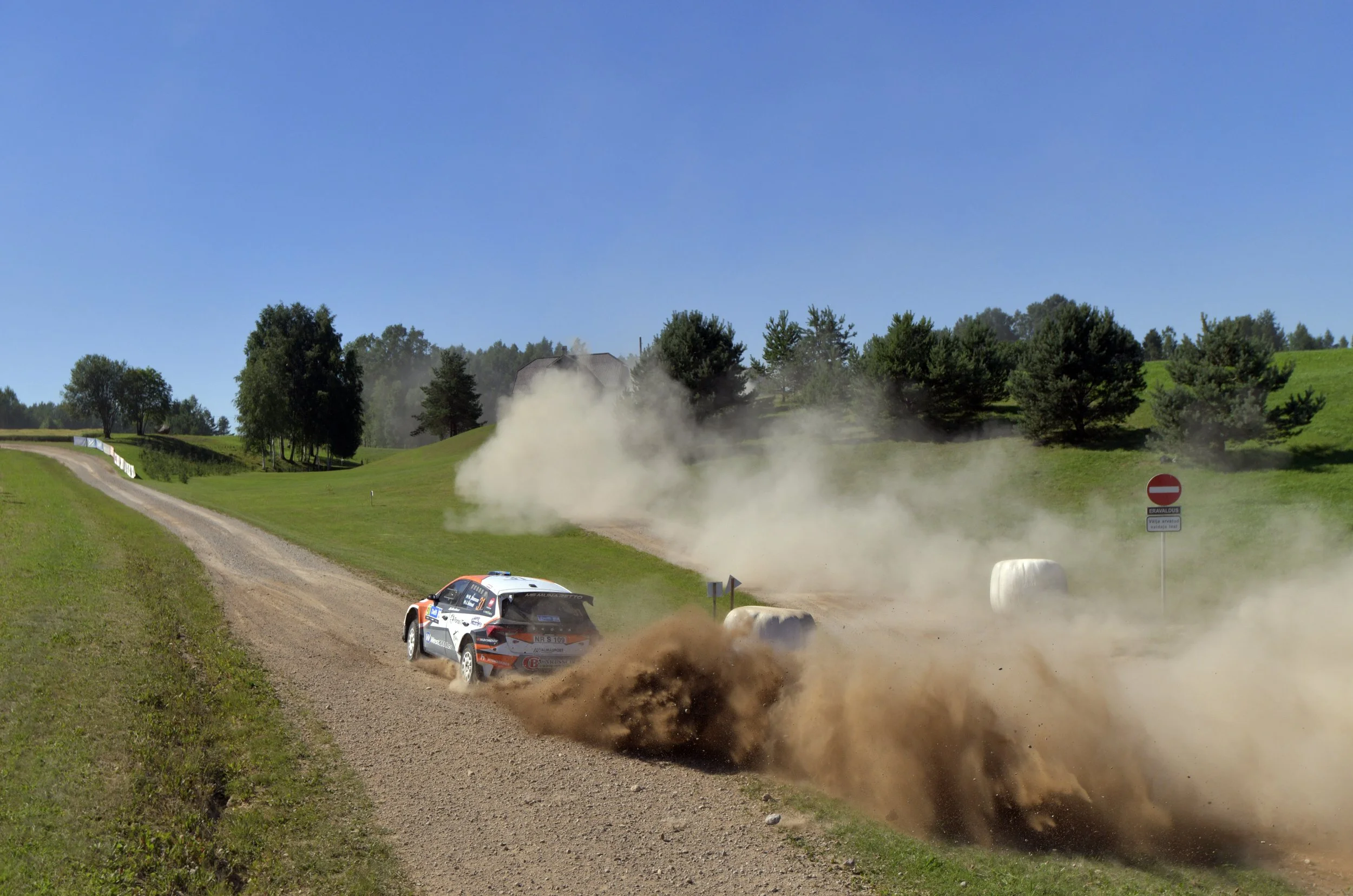 A rally car on a gravel dirt road kicking up dust during a race, with green fields, trees, and a clear blue sky in the background.
