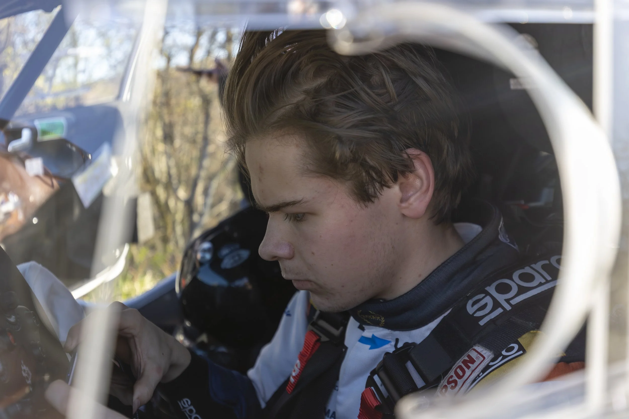 Close-up of a young man with brown hair, wearing a racing suit, sitting inside a vehicle, with trees visible outside through the window.