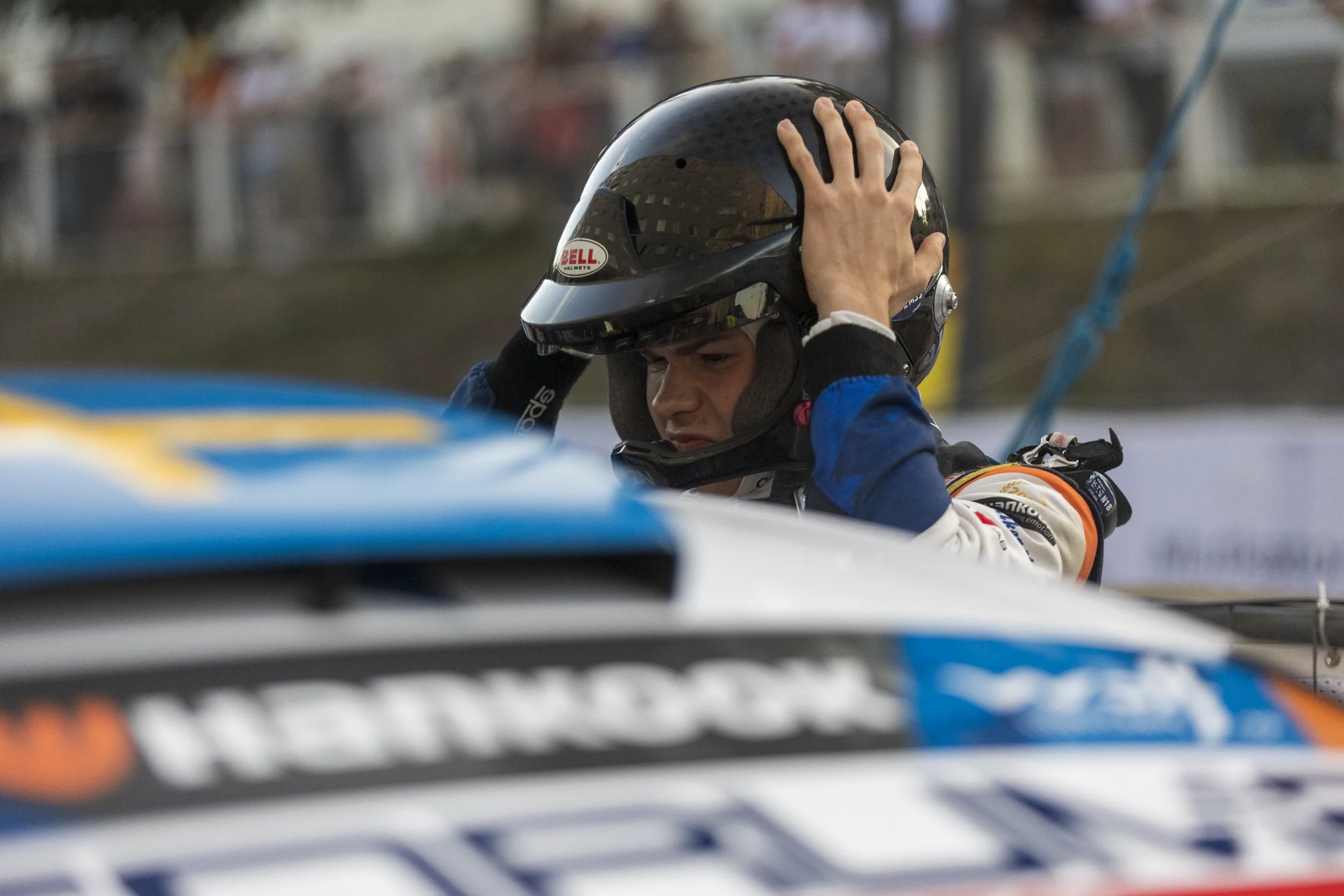 A race car driver wearing a black helmet with a Bell logo, adjusting the helmet strap, with a focused expression on his face. The background is blurred, showing stands and people, indicating a race environment.