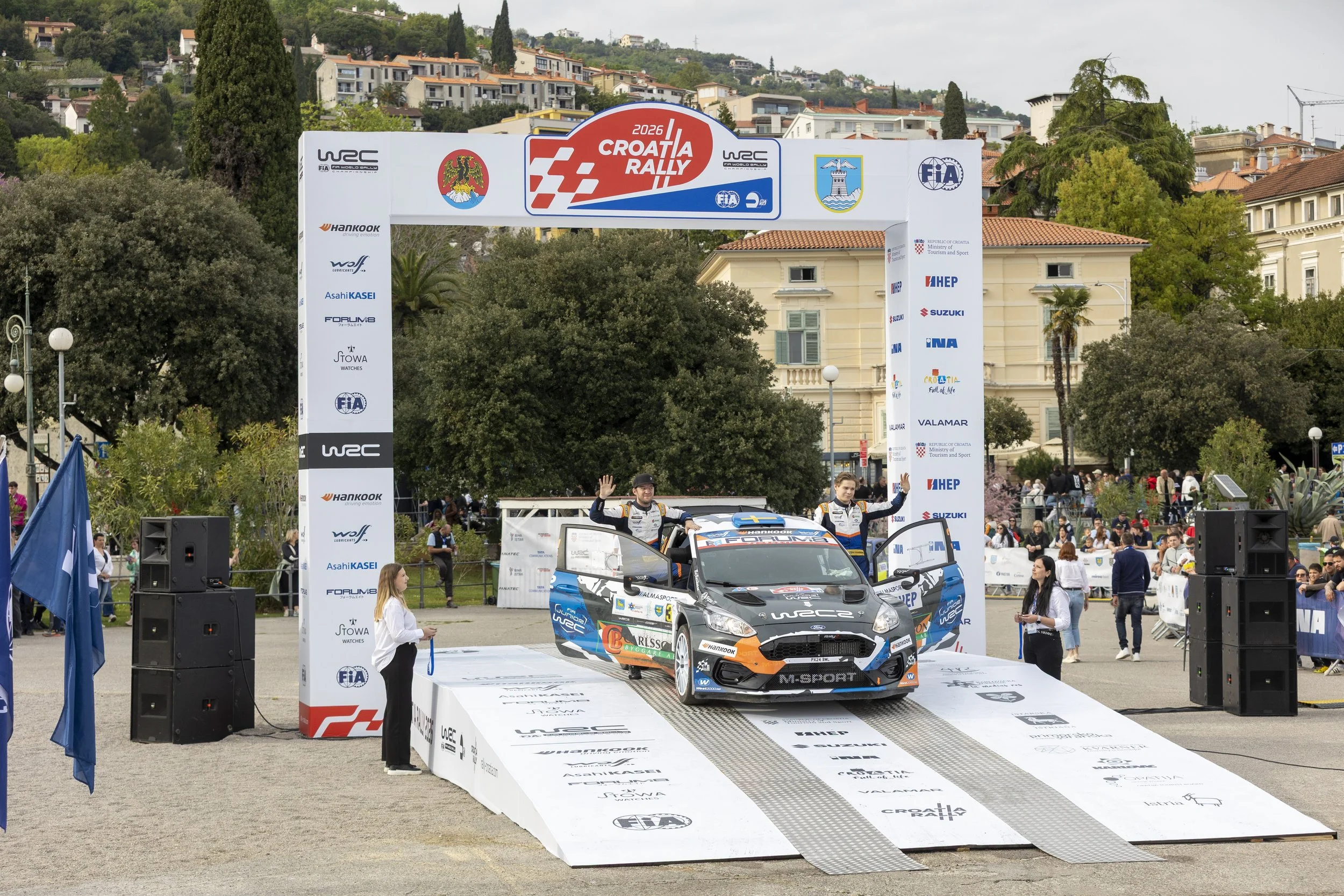 Rally car on podium at Croatia Rally 2026 event with two drivers waving, surrounded by spectators and event banners in an outdoor city setting.