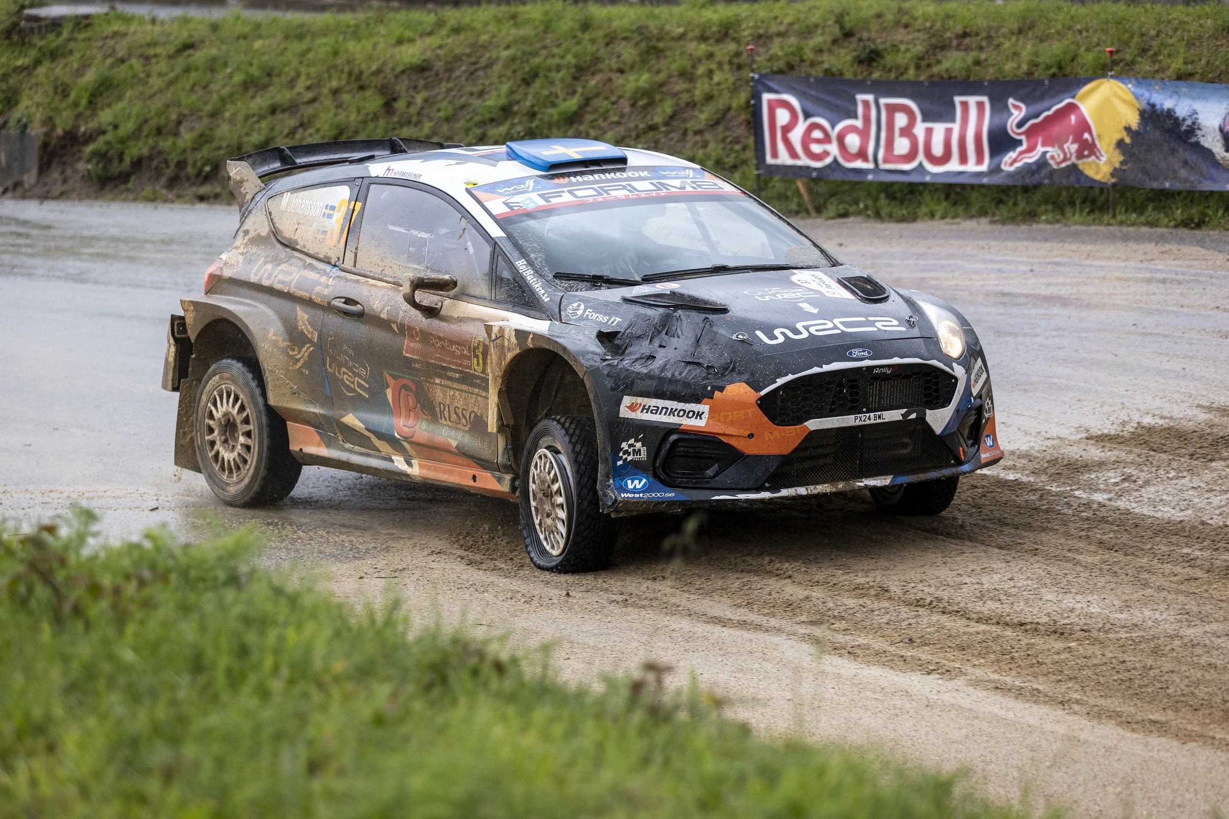 A black rally car driving on a wet dirt road, covered in mud, with a Red Bull banner in the background.