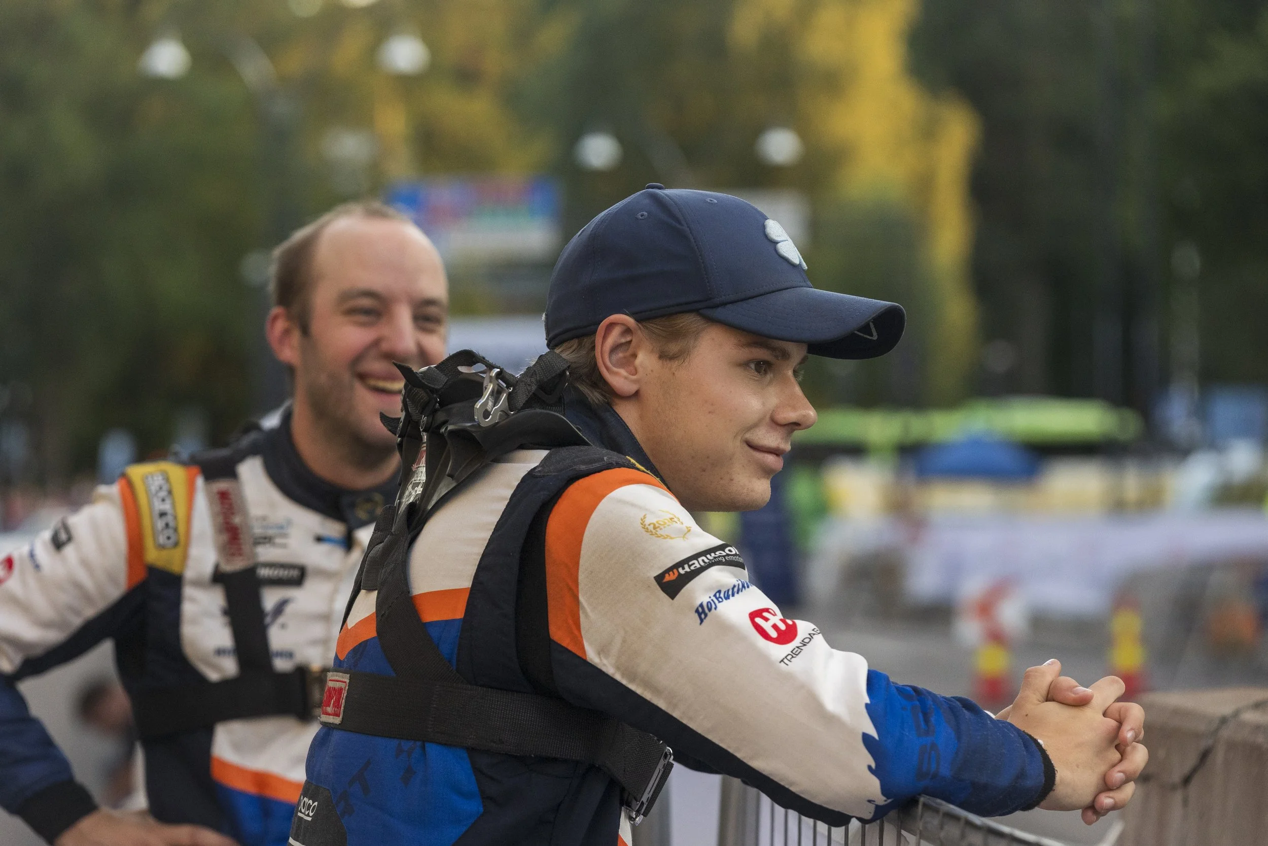 Two race car drivers in racing suits and helmets standing behind a metal barrier, smiling and chatting at a racing event with blurred trees and structures in the background.