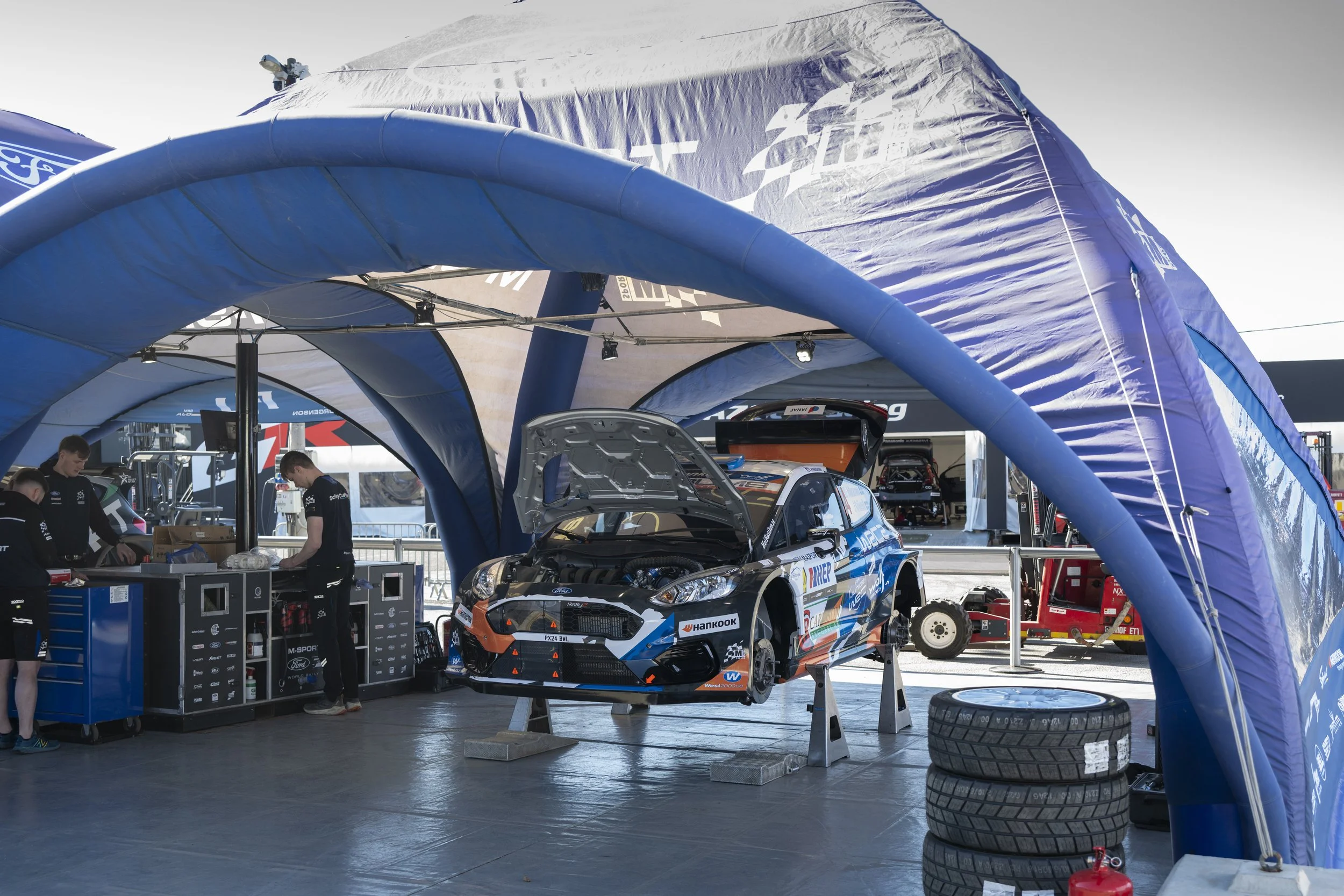 View of a racing car inside a pit area under a blue tent, with team members working, tools, spare tires, and various equipment visible.