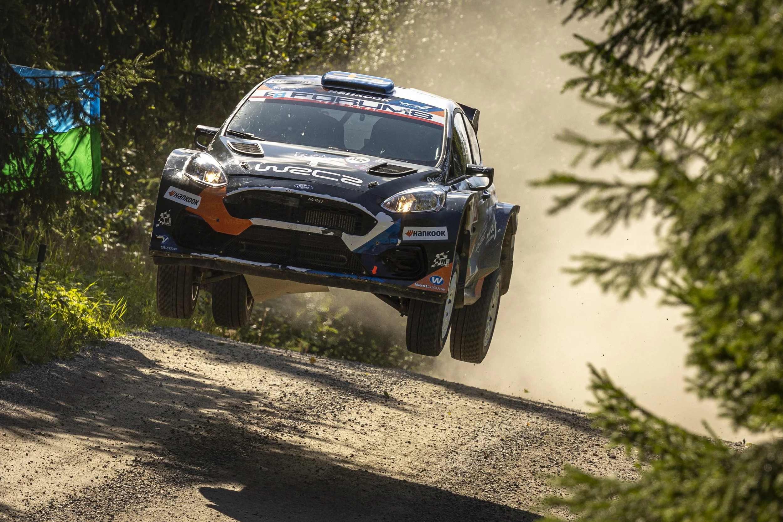 A rally car flying through the air on a dirt rally stage surrounded by trees.