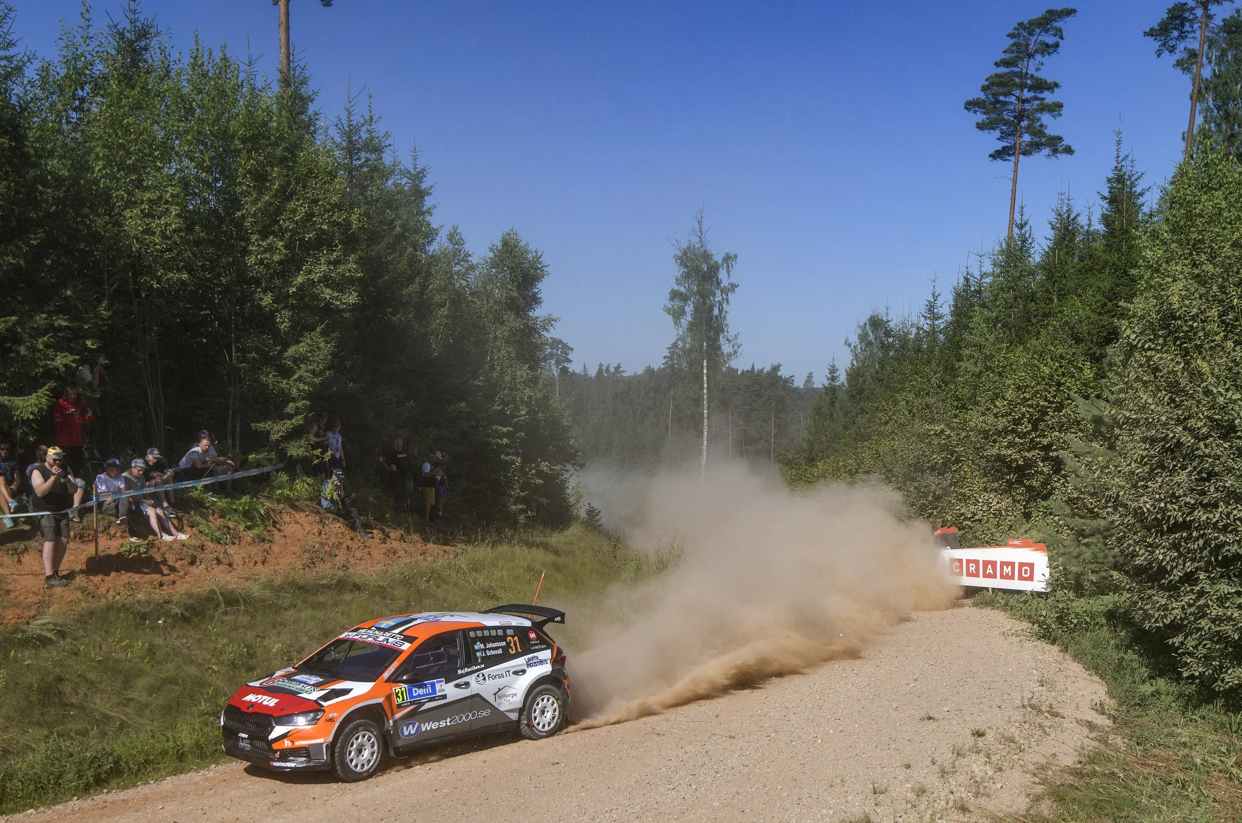 A rally car racing on a dirt road in a forest, with spectators watching from the side. The car is kicking up dust as it speeds around a corner, surrounded by tall trees and blue sky.