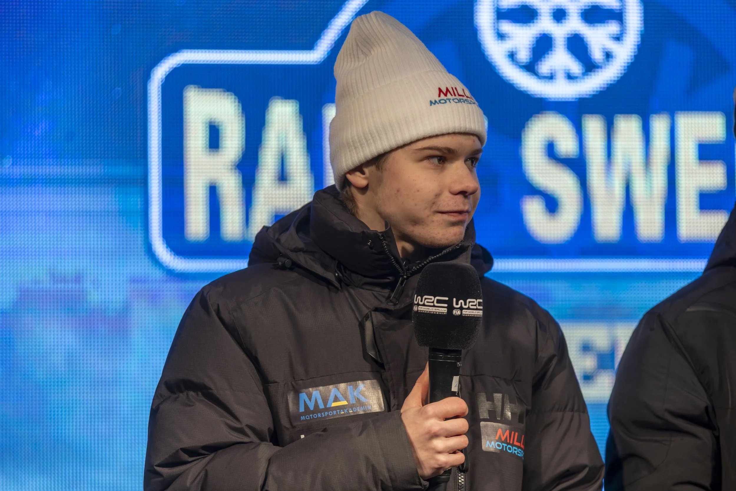 A young man wearing a white beanie and a dark jacket holding a microphone with a "WRC" logo, during an interview or press event with a digital blue background displaying the words "Race" and other text.