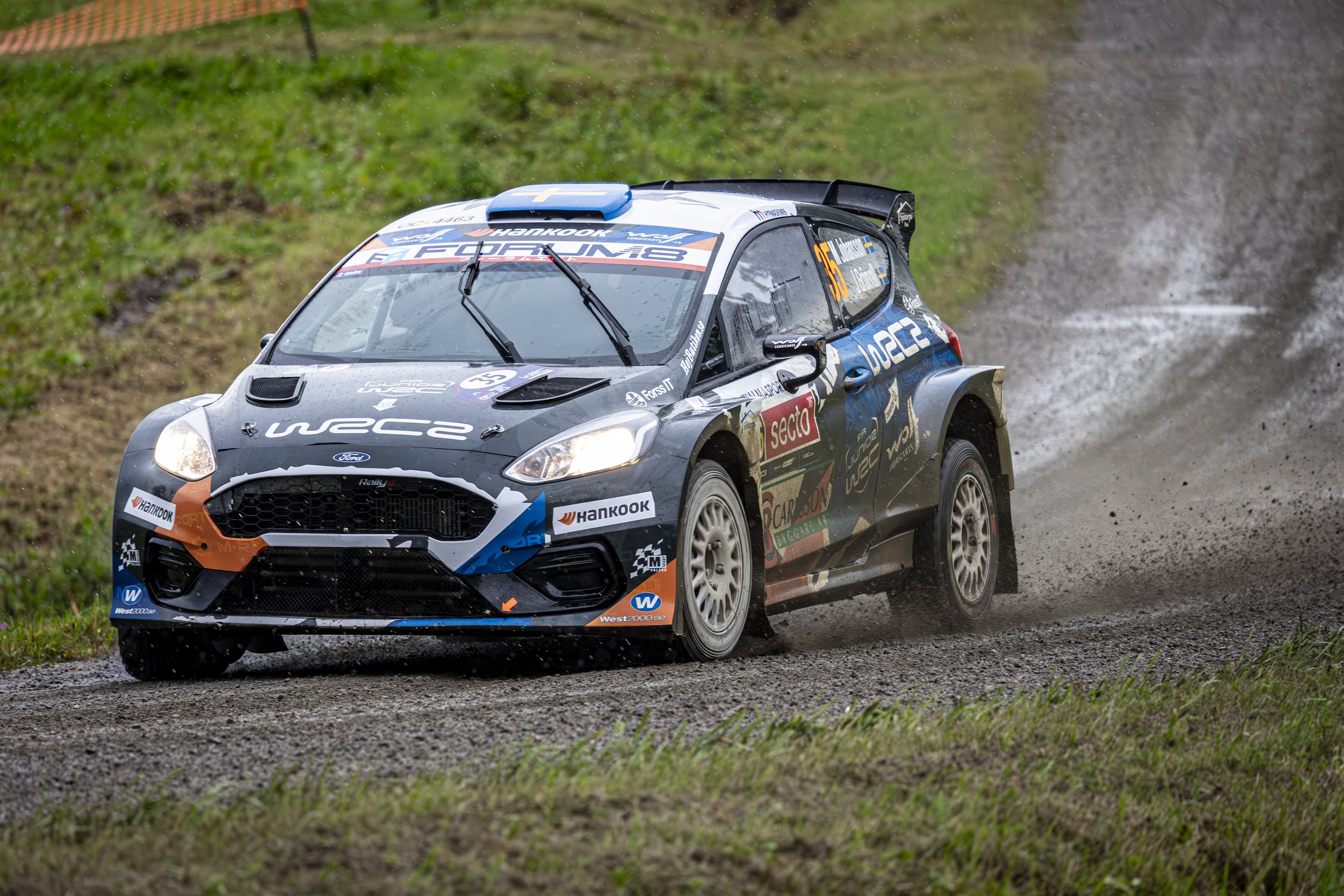 A rally car speeding through a muddy, wet rally stage, kicking up water and mud as it moves along a gravel road with green grass and foliage on either side.