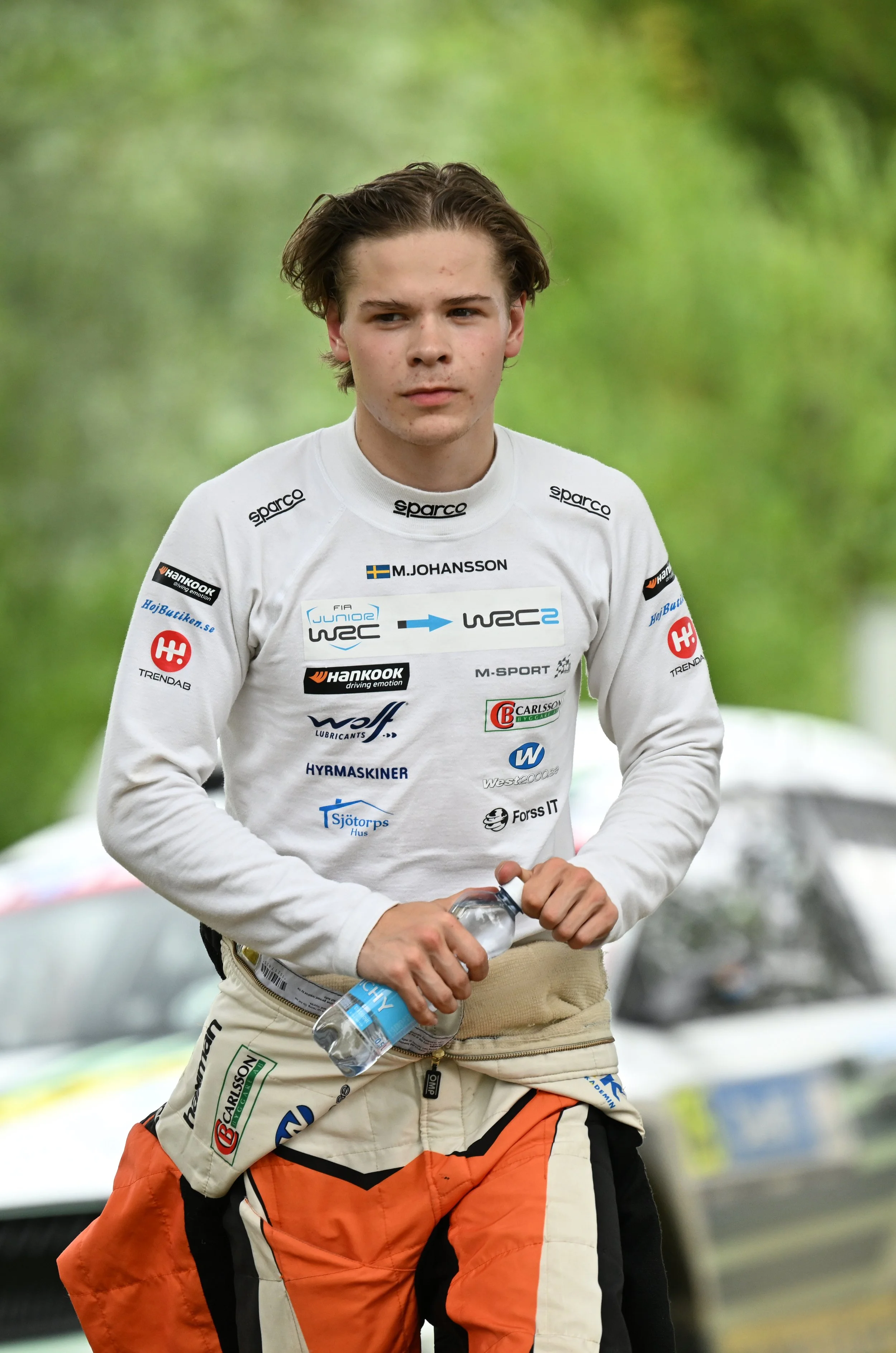 A young male race car driver holding a water bottle and wearing a racing suit with sponsor logos, walking outdoors with green trees in the background.