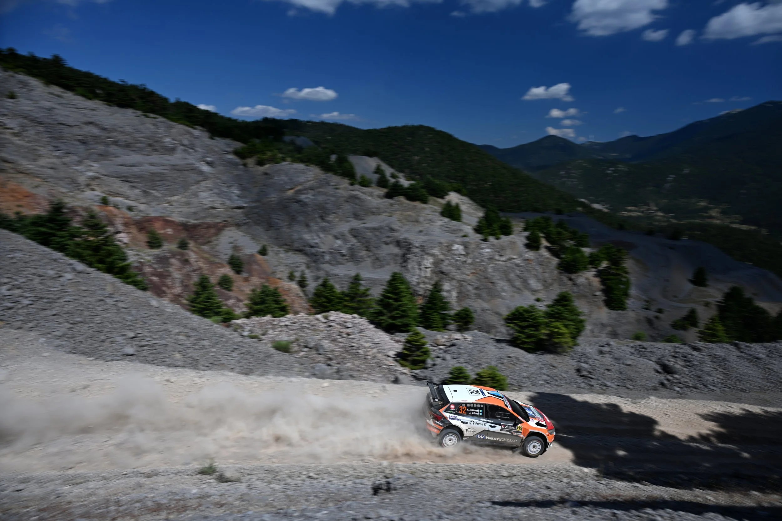 Rally car racing on a dirt mountain road with mountain scenery and blue sky in the background.