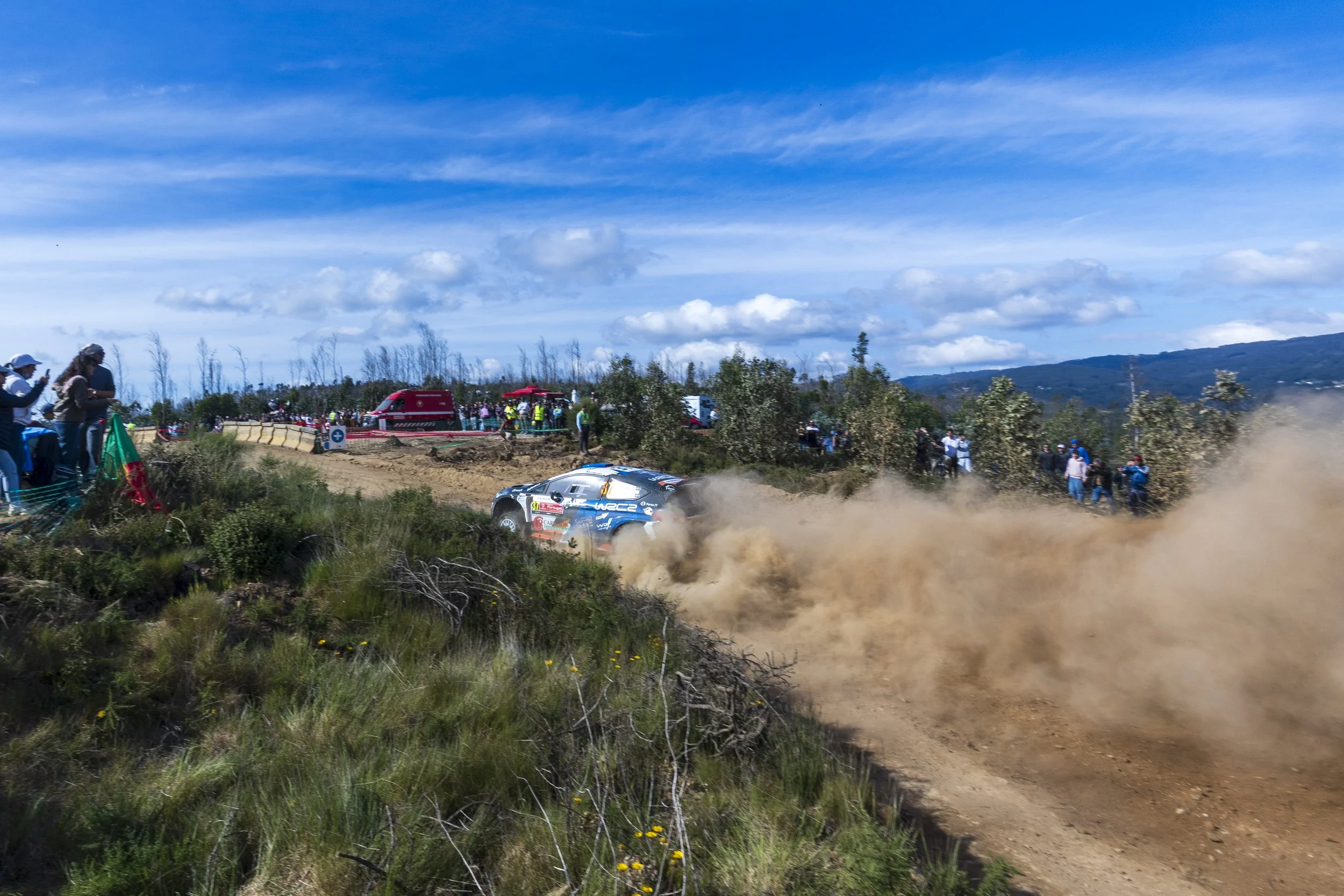 A rally car speeding on a dirt track, kicking up dust, with spectators watching nearby under a partly cloudy sky.