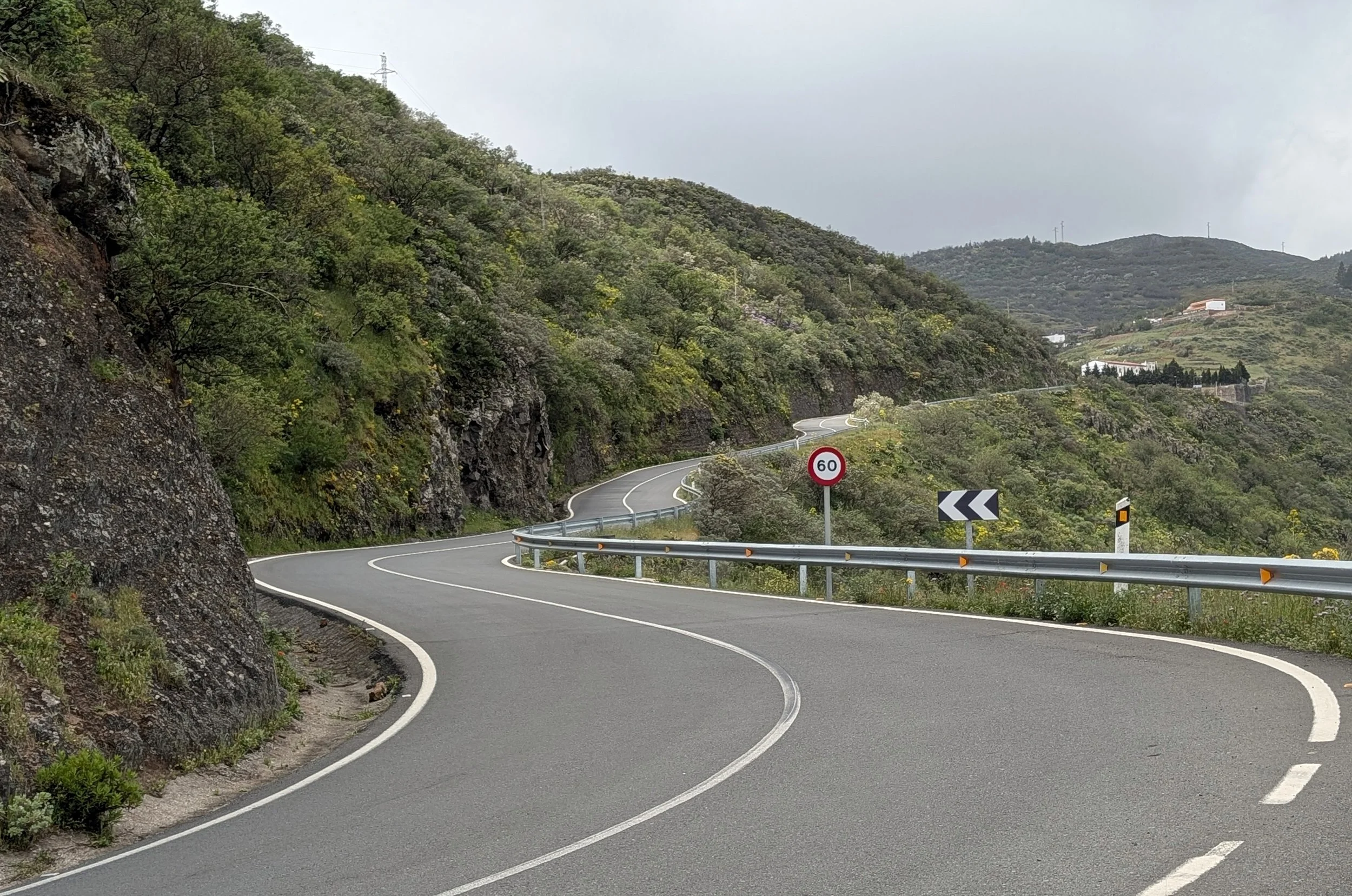 A winding mountain road with guardrails, surrounded by green hills and trees, under a cloudy sky, with a speed limit sign showing 60.