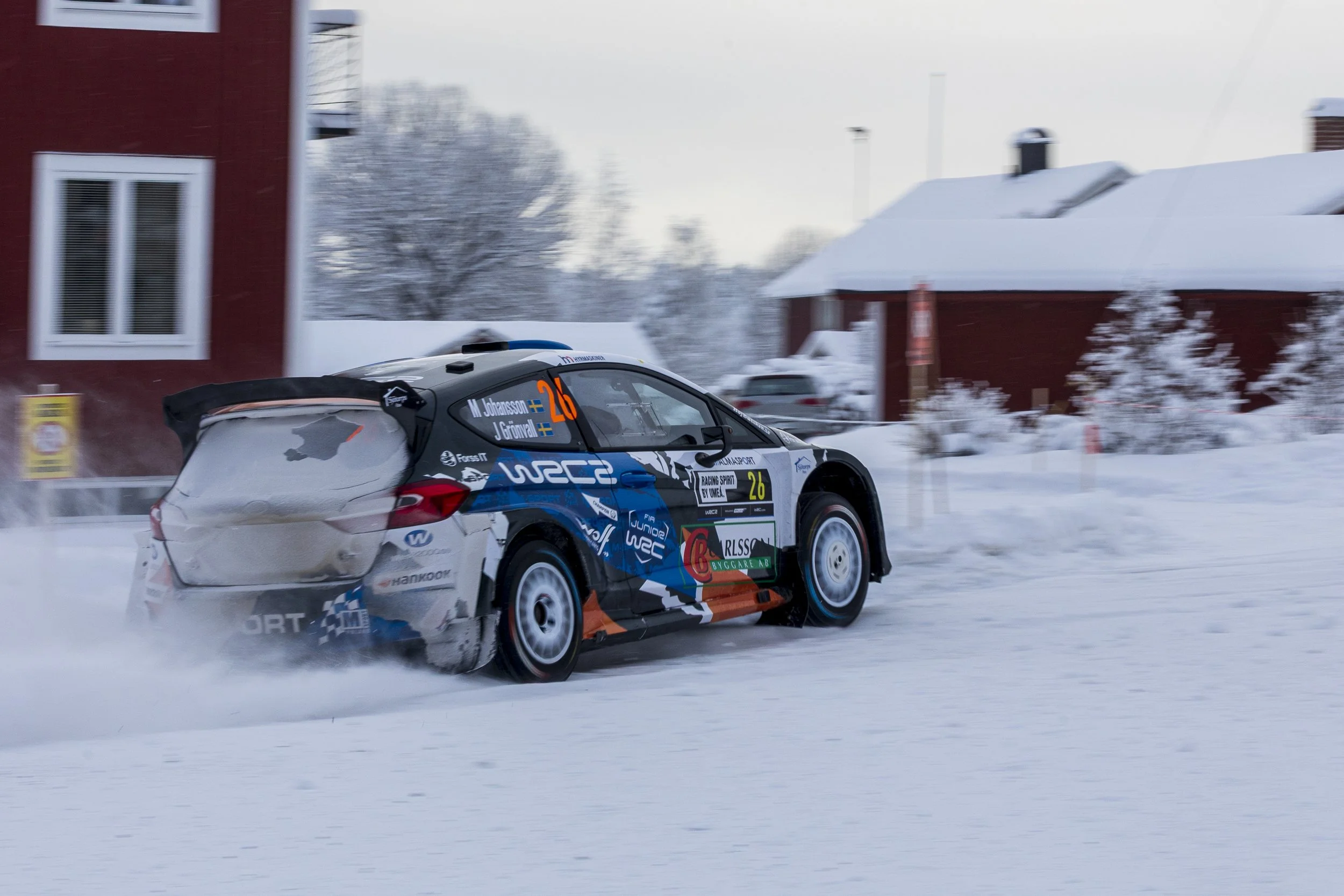 A rally car racing on a snowy road during winter, with snow-covered buildings and trees in the background.