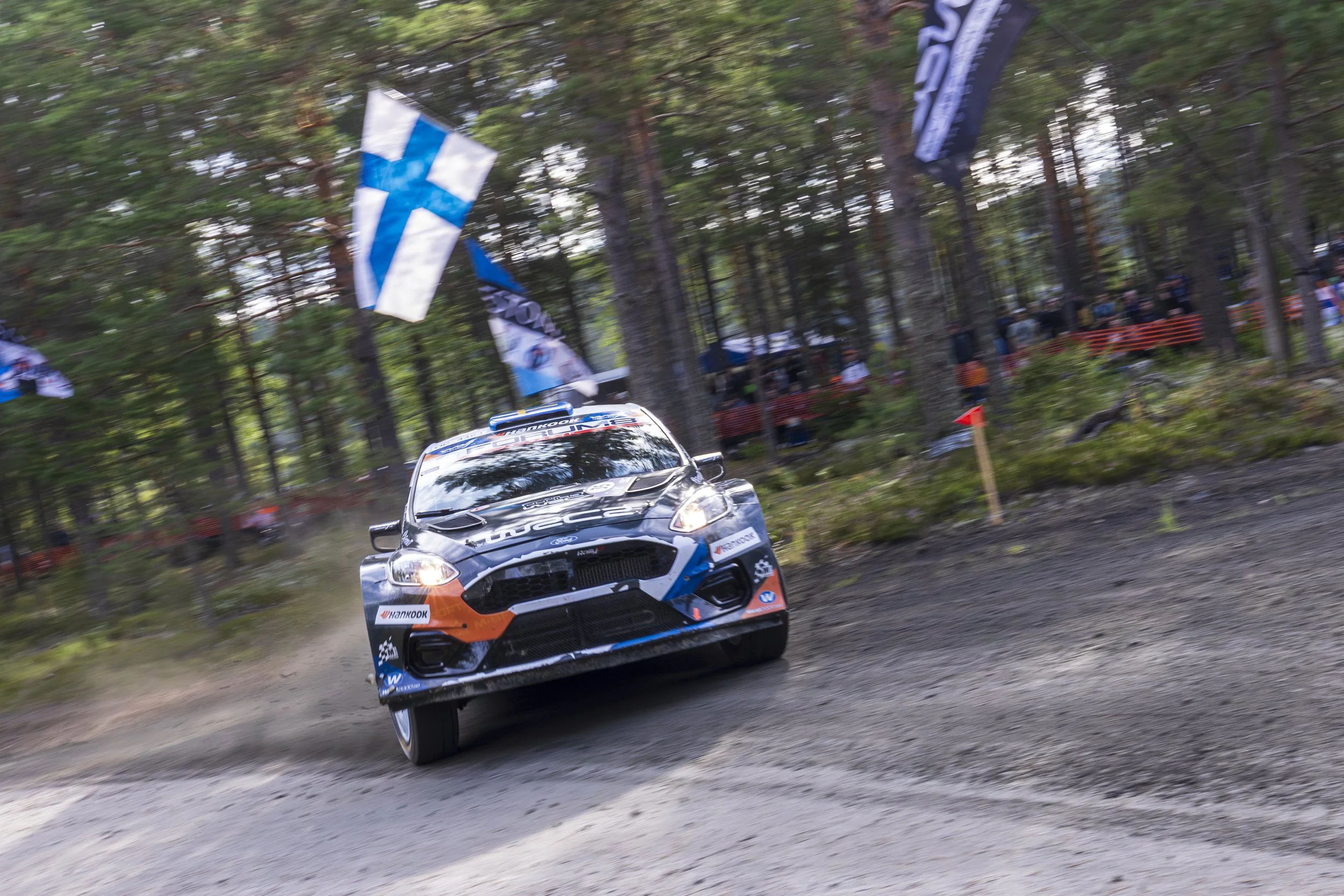 A rally car speeding on a dirt road through a forest, with flags and spectators in the background.
