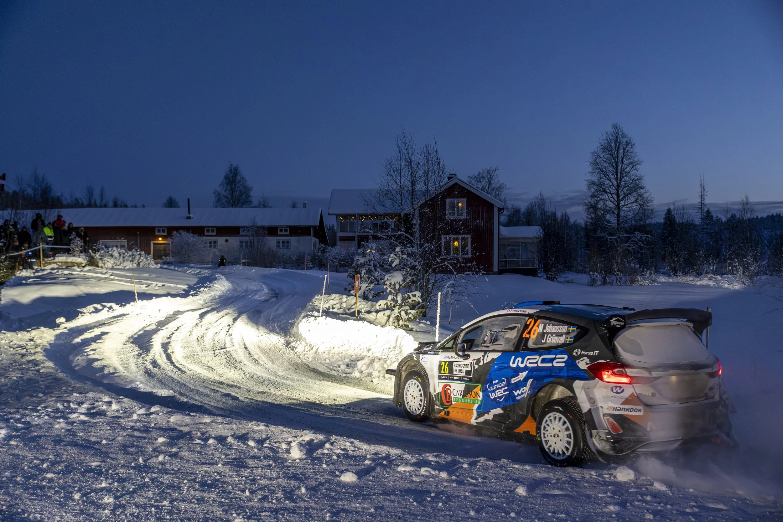 A rally car on a snow-covered race track during dusk with houses and trees in the background, and a group of people watching.