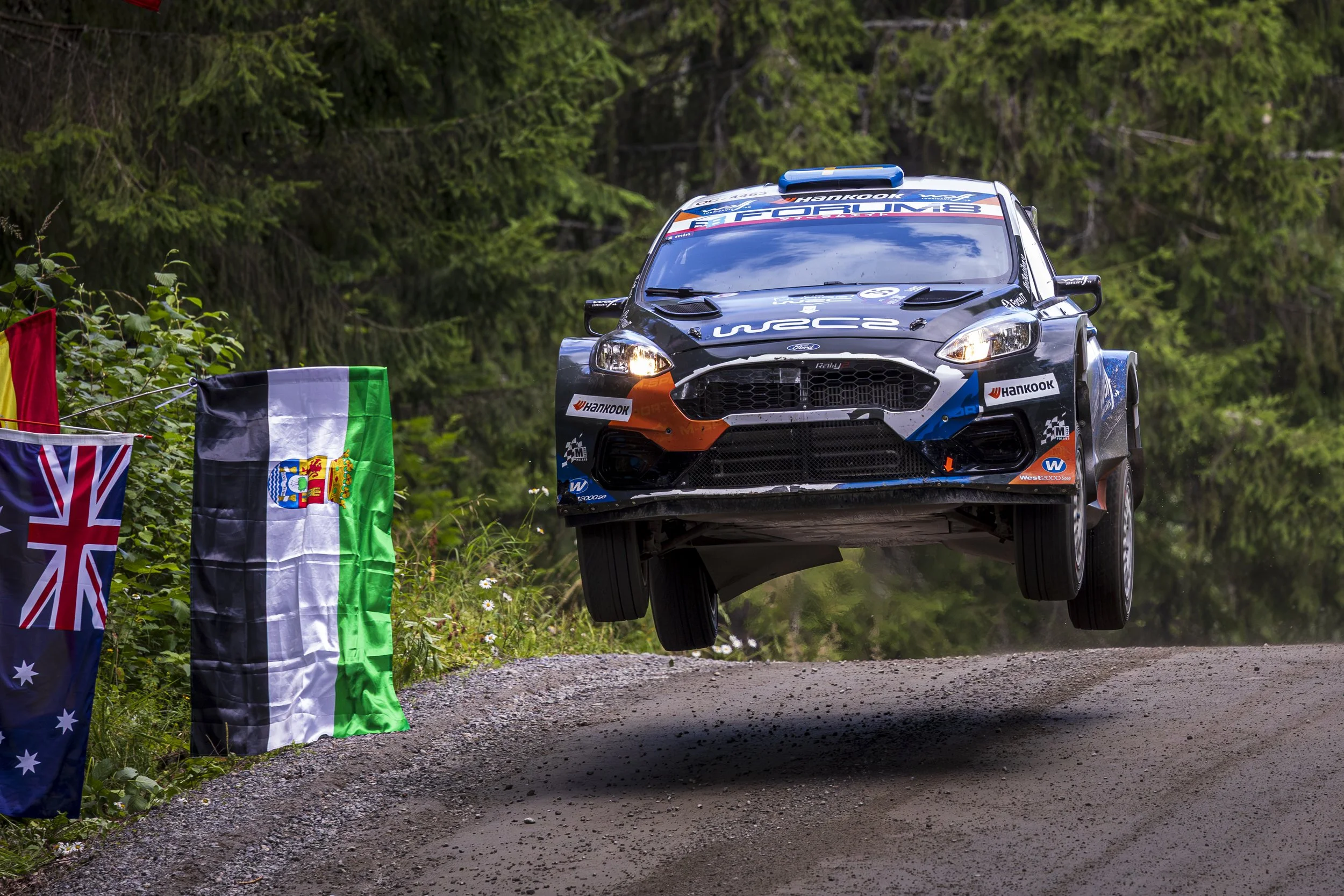 A black rally car with sponsors and decals, mid-air during a rally race on a dirt road surrounded by greenery and trees, with flags hanging on the side.