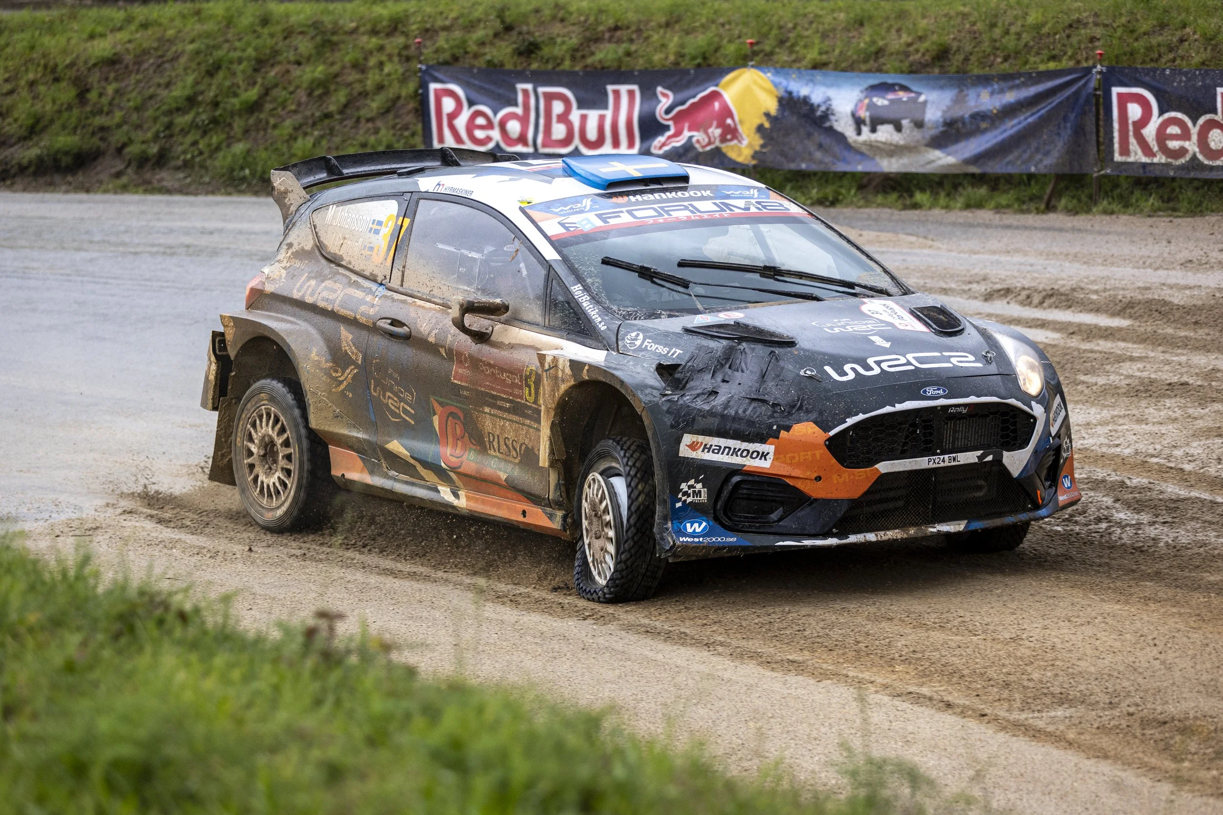 A rally car speeding on a dirt track with mud splashed on its body, featuring a "WRC" logo on the hood and various sponsor logos. The background includes a Red Bull banner along the grassy hillside.