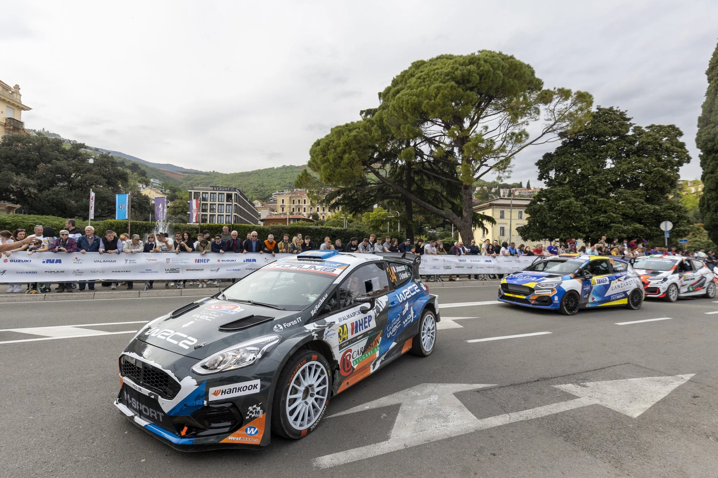 Rally cars racing with spectators behind safety barriers in an urban setting with buildings and green hills in the background.