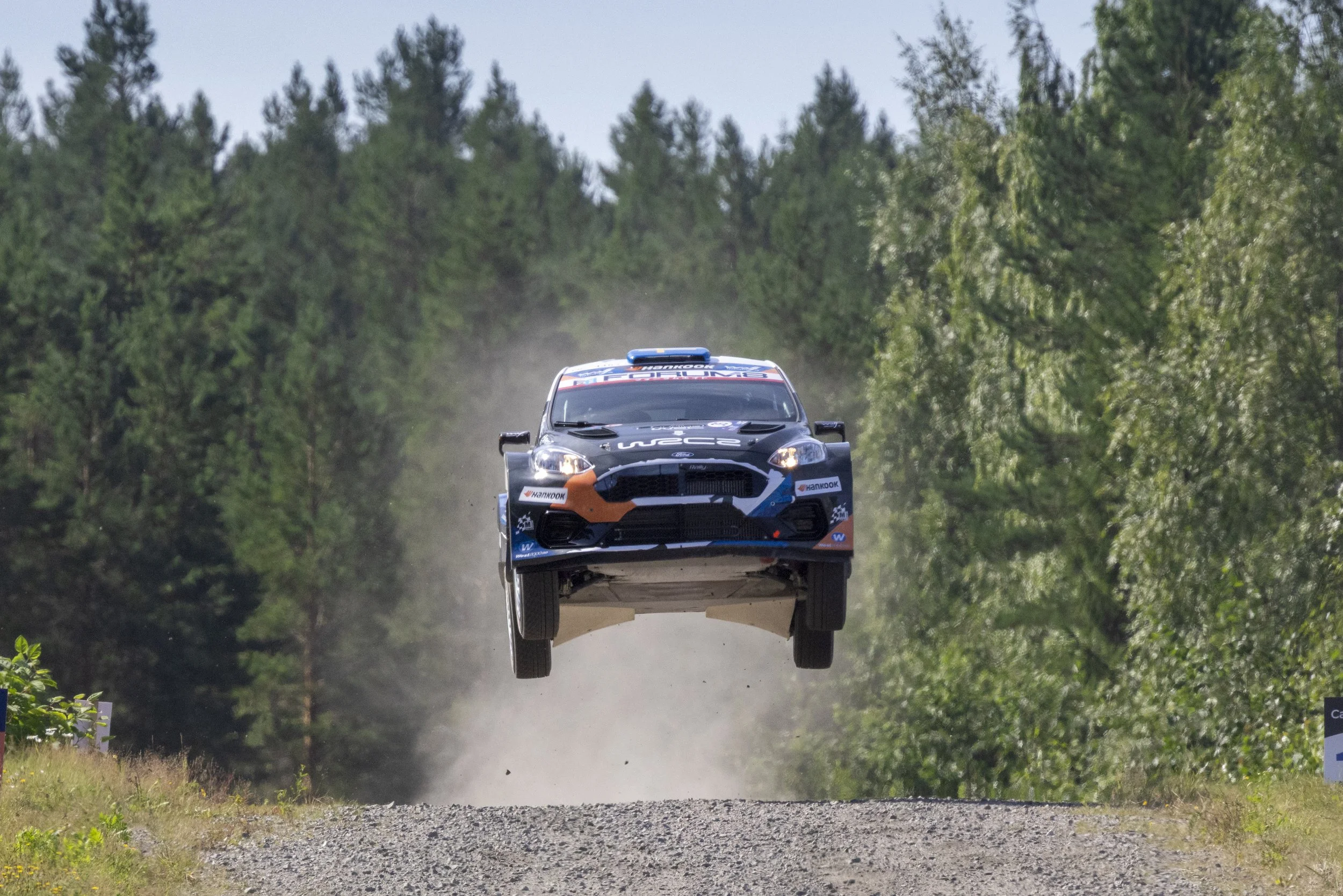 A rally car flying through the air over a dirt road surrounded by a forest.