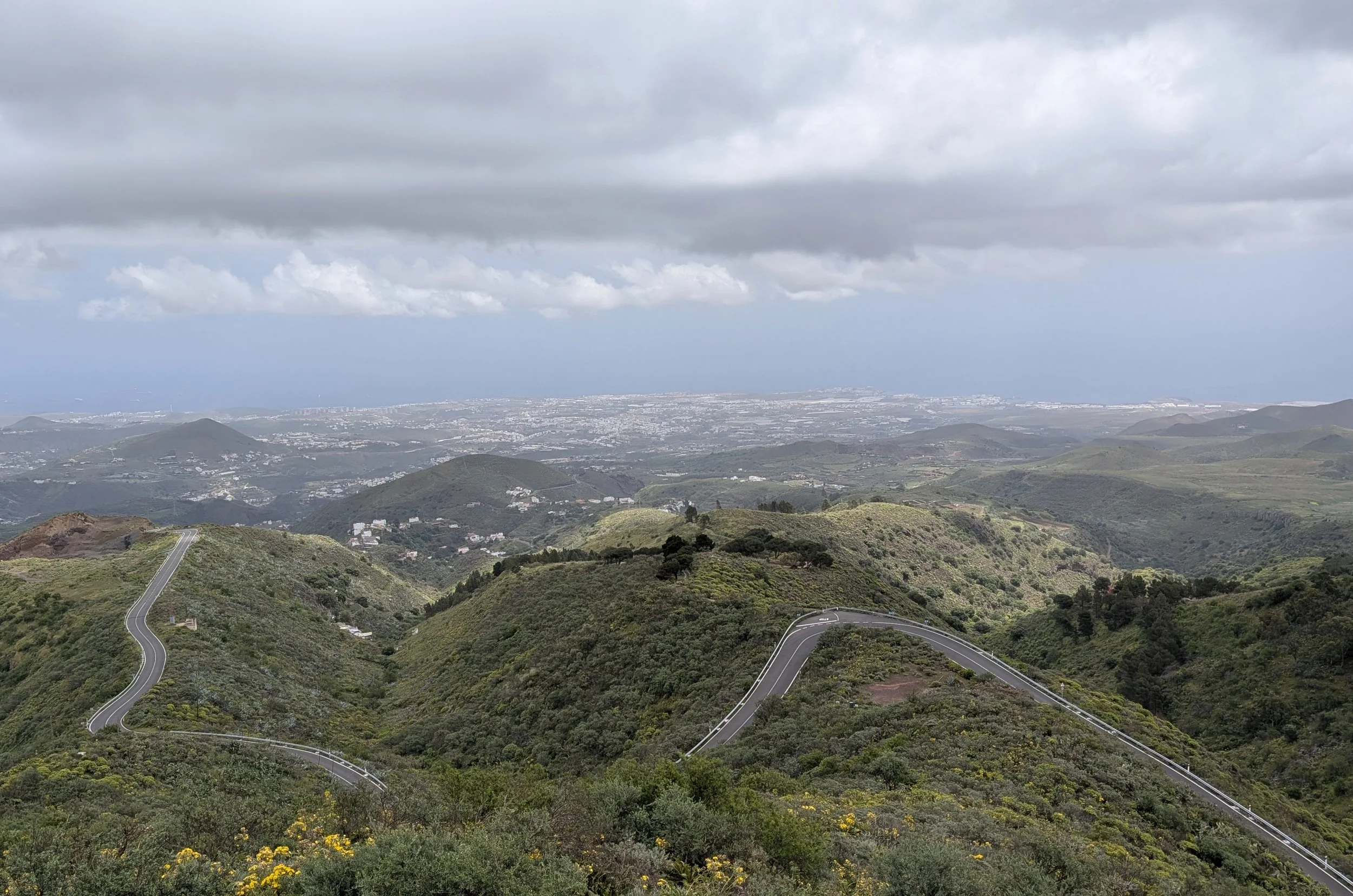 Scenic view of winding mountain roads through green hills with distant cityscape and cloudy sky overhead.