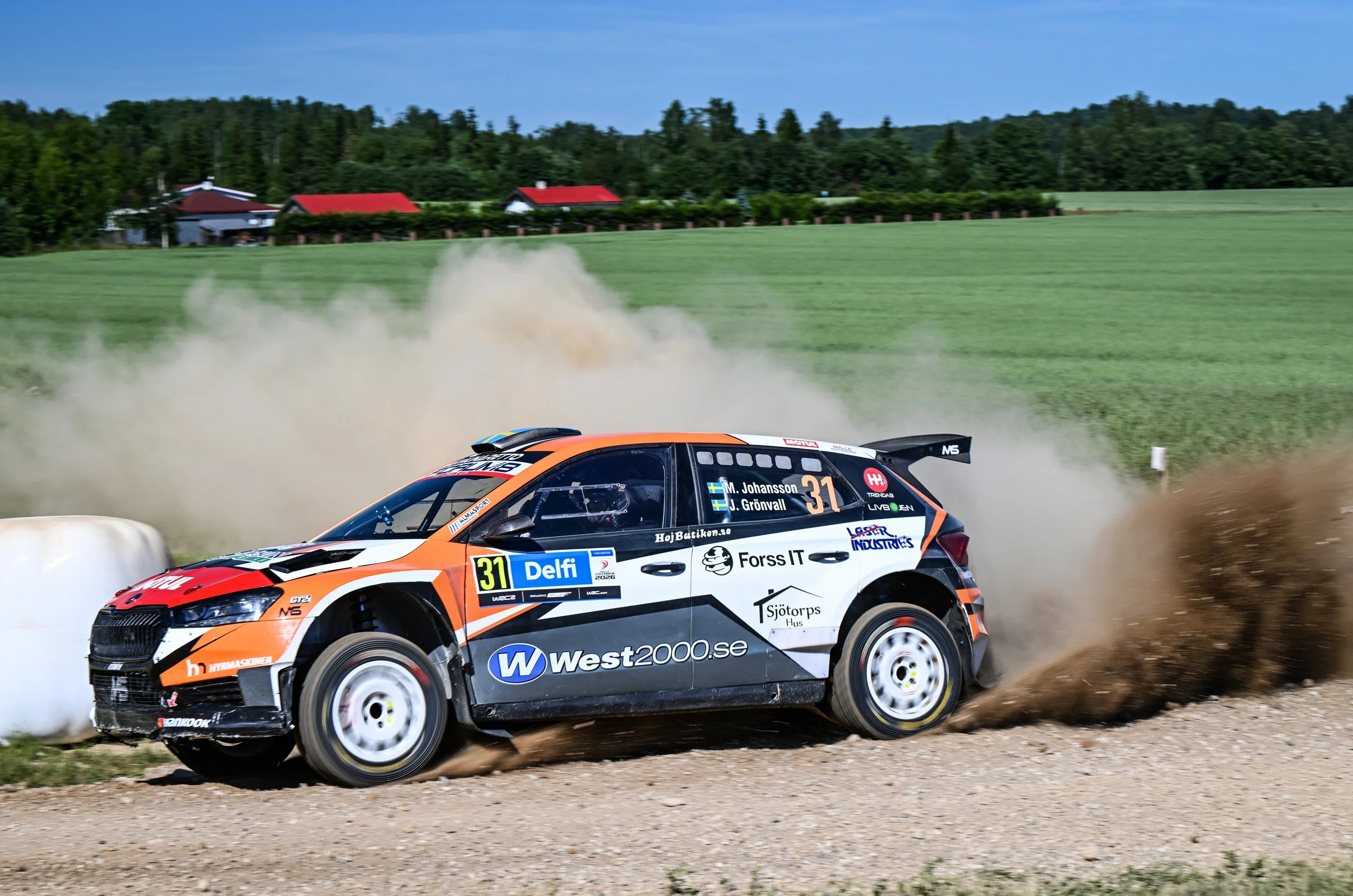 A rally car racing on a gravel track kicks up dust, with green fields and trees in the background.