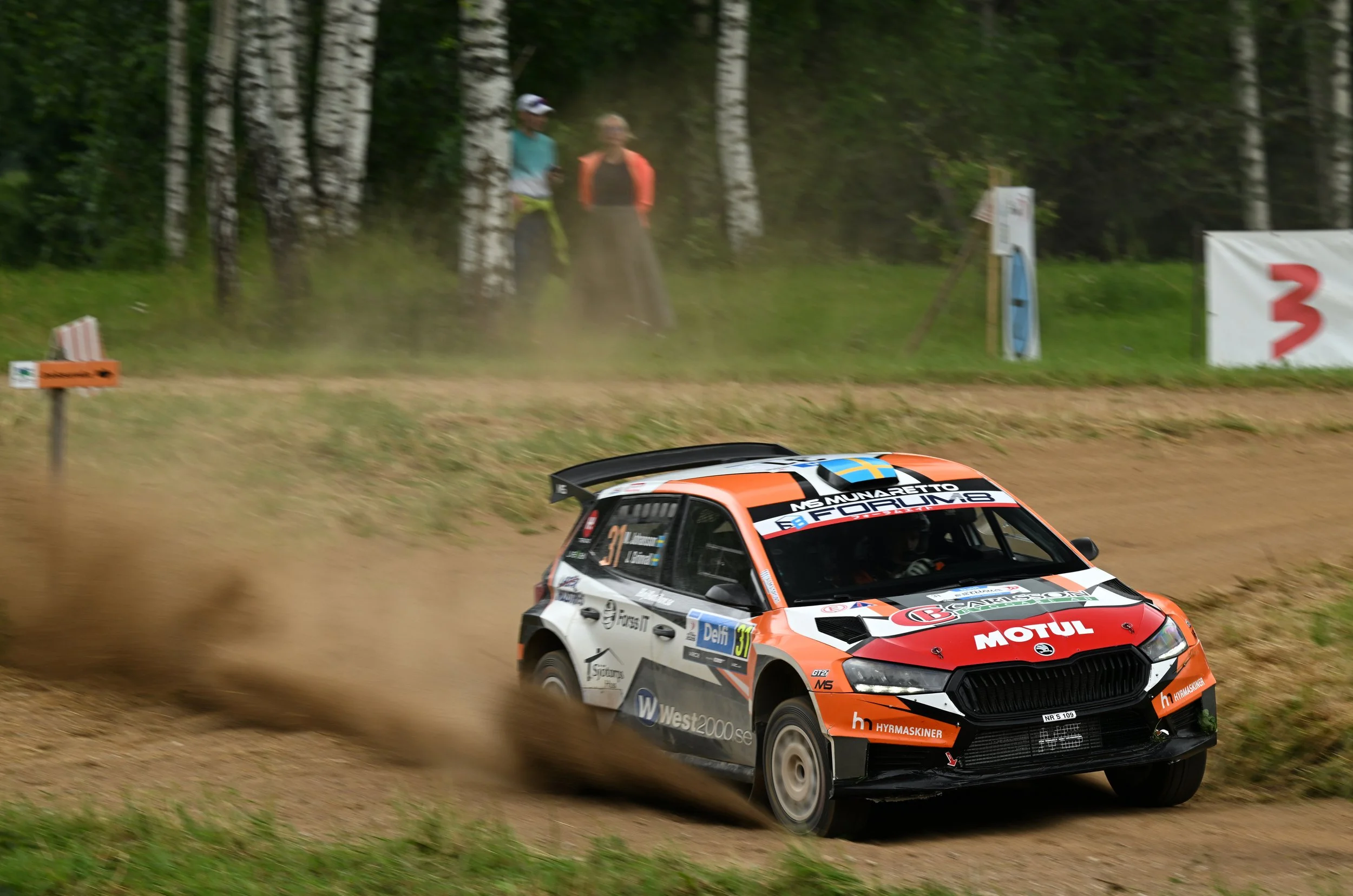 A rally car driving on dirt road, kicking up dust, with a forested background and two spectators watching from the side.