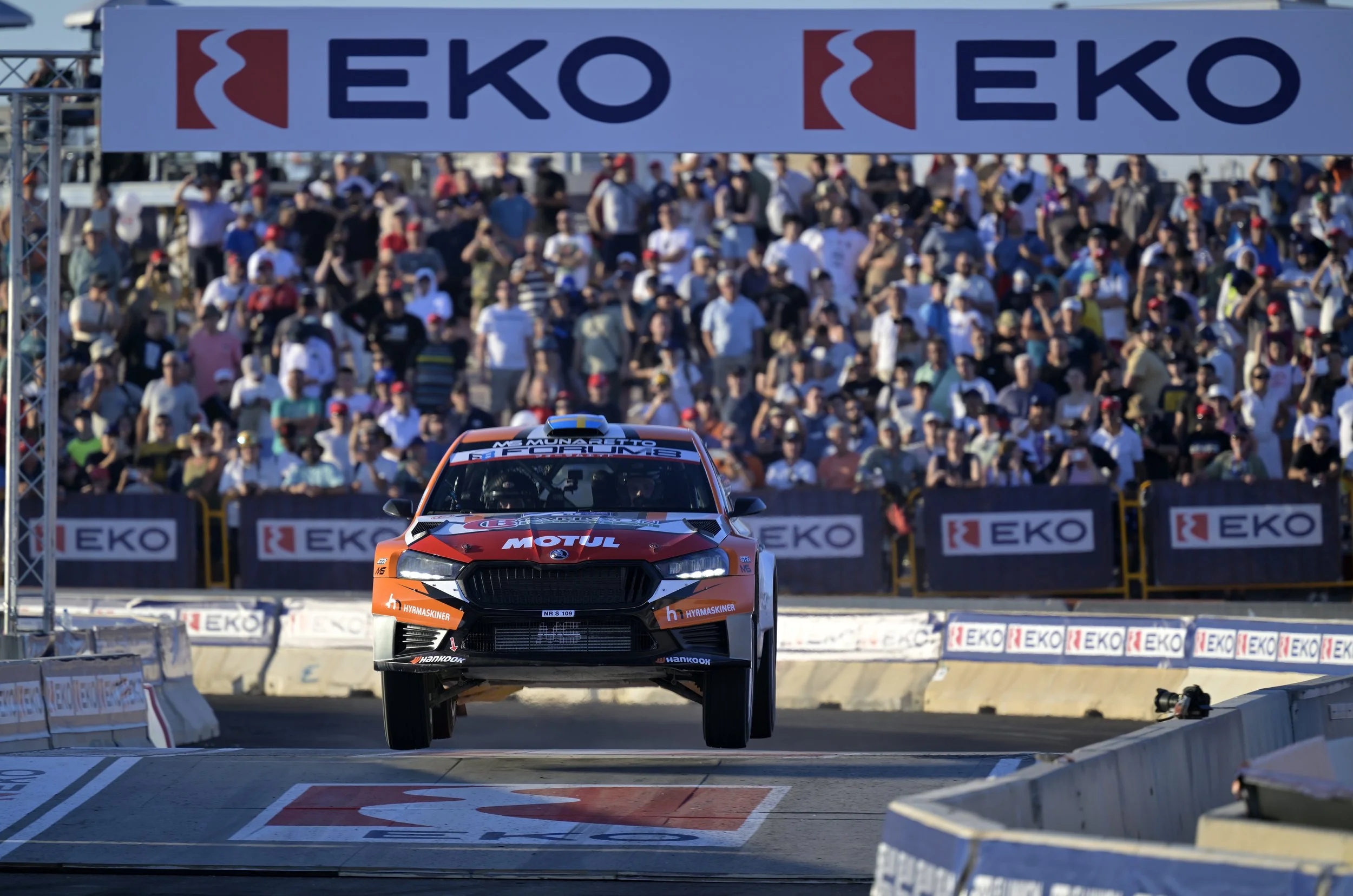 A race car is airborne and jumping off a ramp during a motorsports event with a large crowd of spectators in the background.