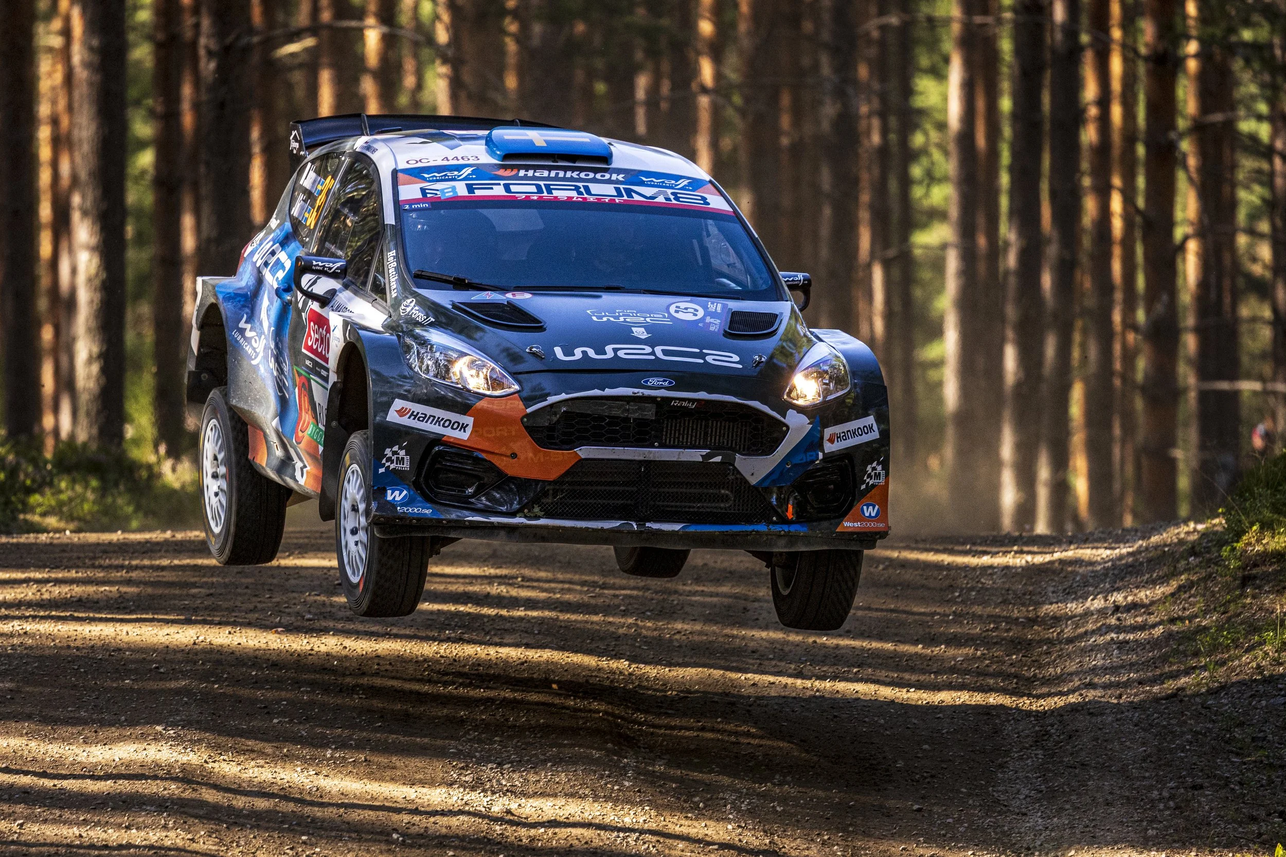 A blue rally car with sponsor logos driving through a dirt forest trail surrounded by tall trees.